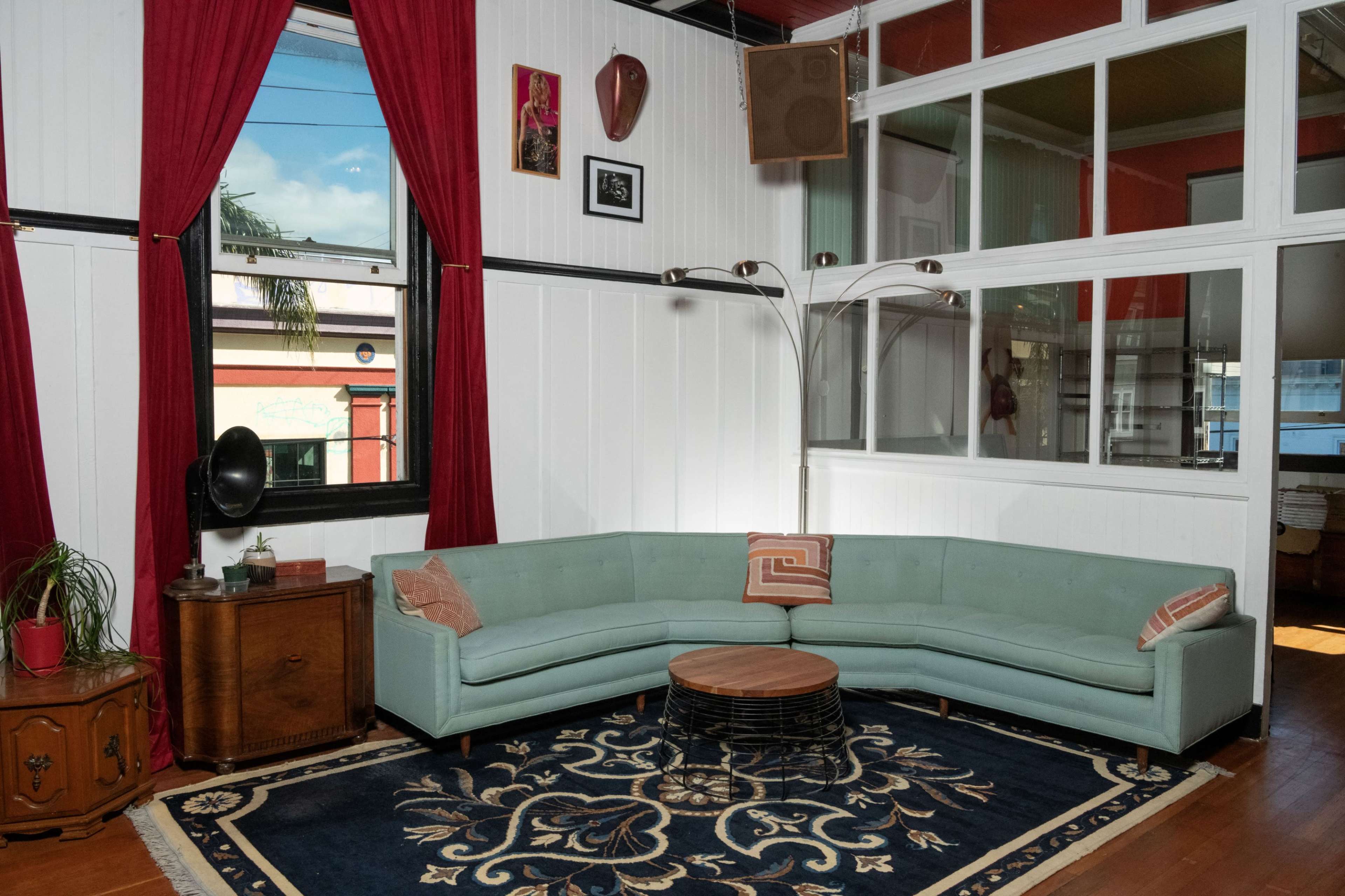 A cozy living room featuring a curved mint green sofa, a round wooden coffee table, a vintage record player, and large windows with red curtains.