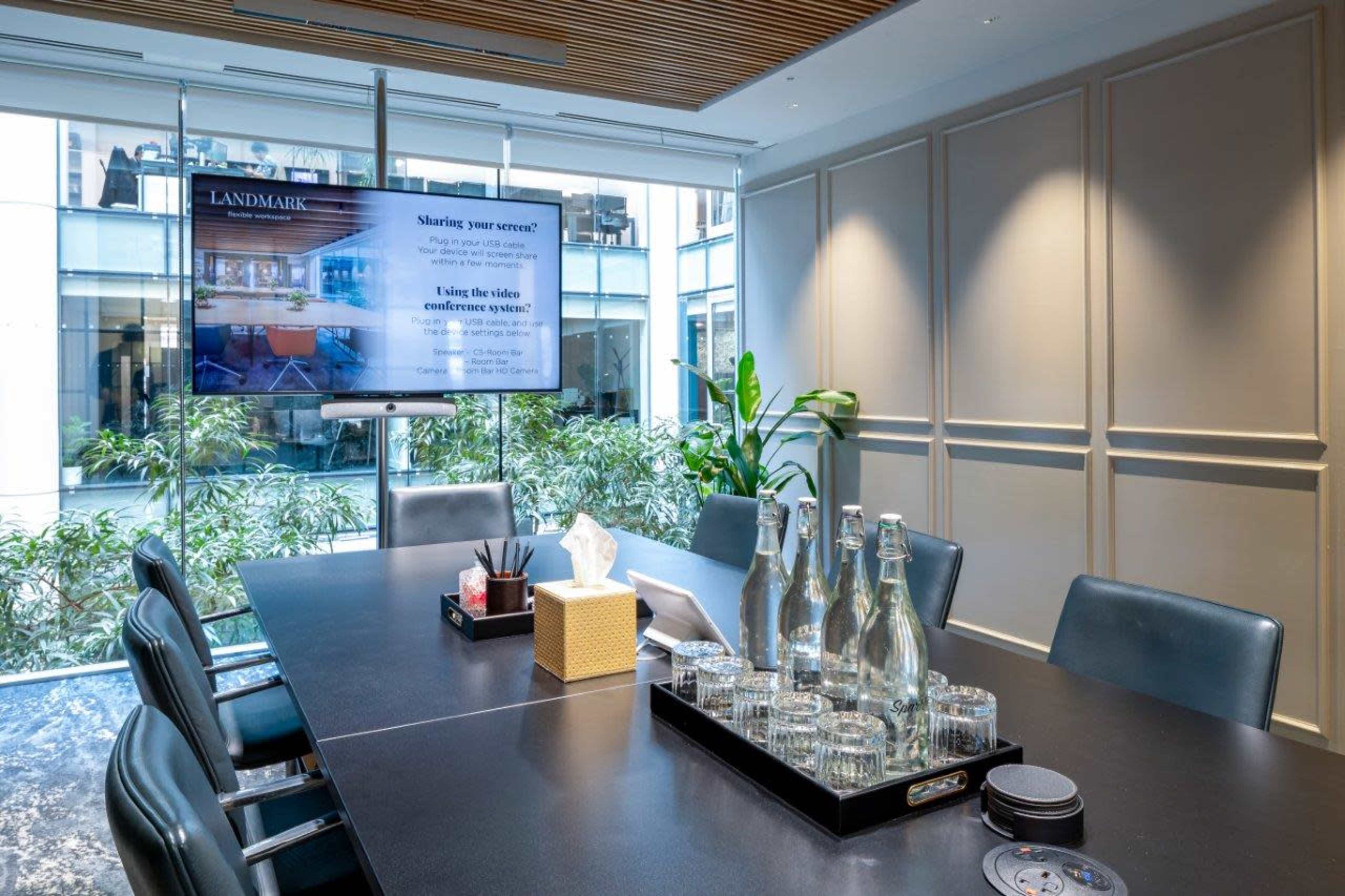 A modern conference room features a large table, glasses, bottled water, and a screen displaying information, surrounded by large windows with greenery outside.