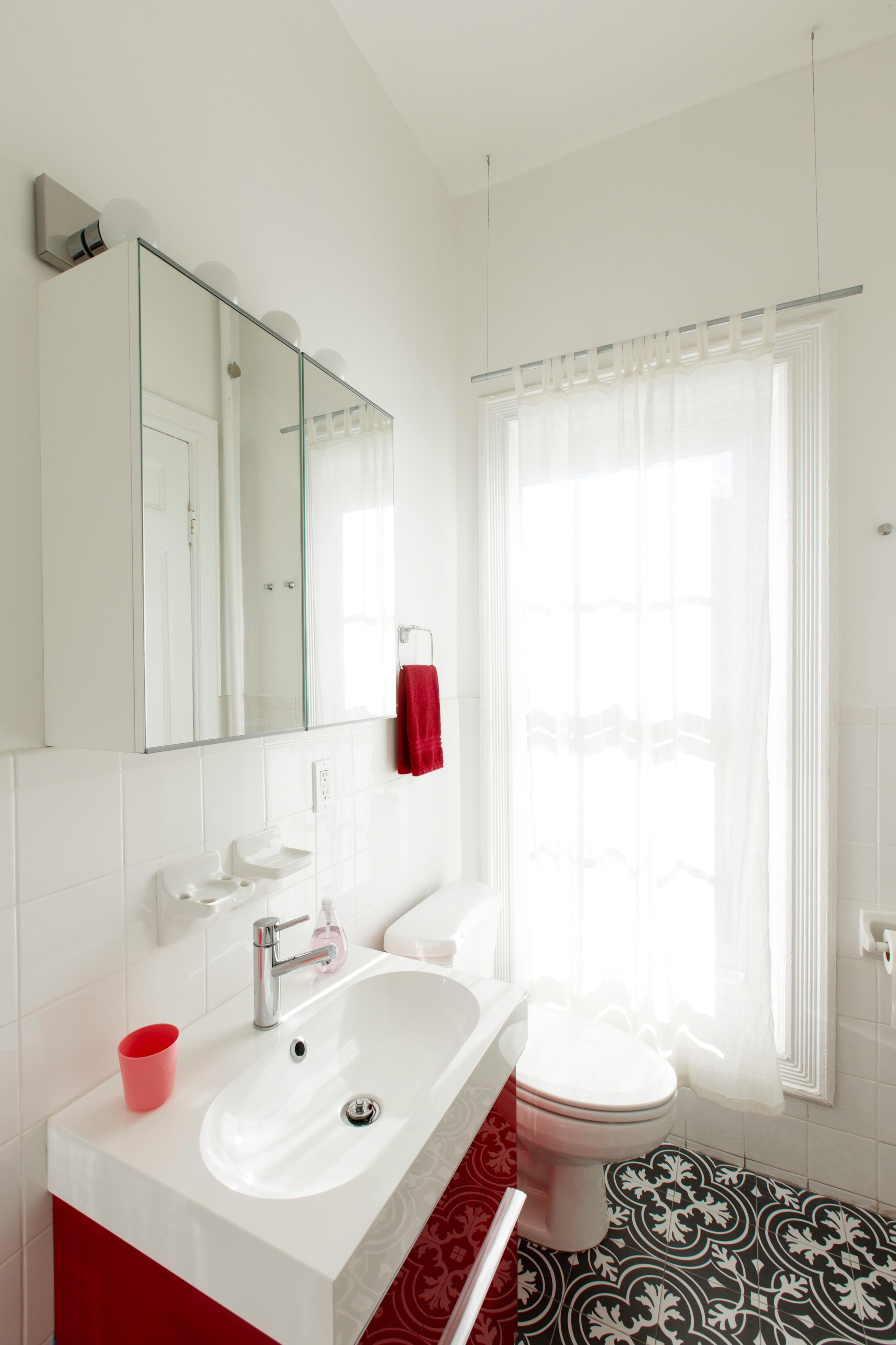 A modern bathroom with a white sink, red cabinetry, a wall-mounted mirror, and a window partially covered by a sheer curtain.