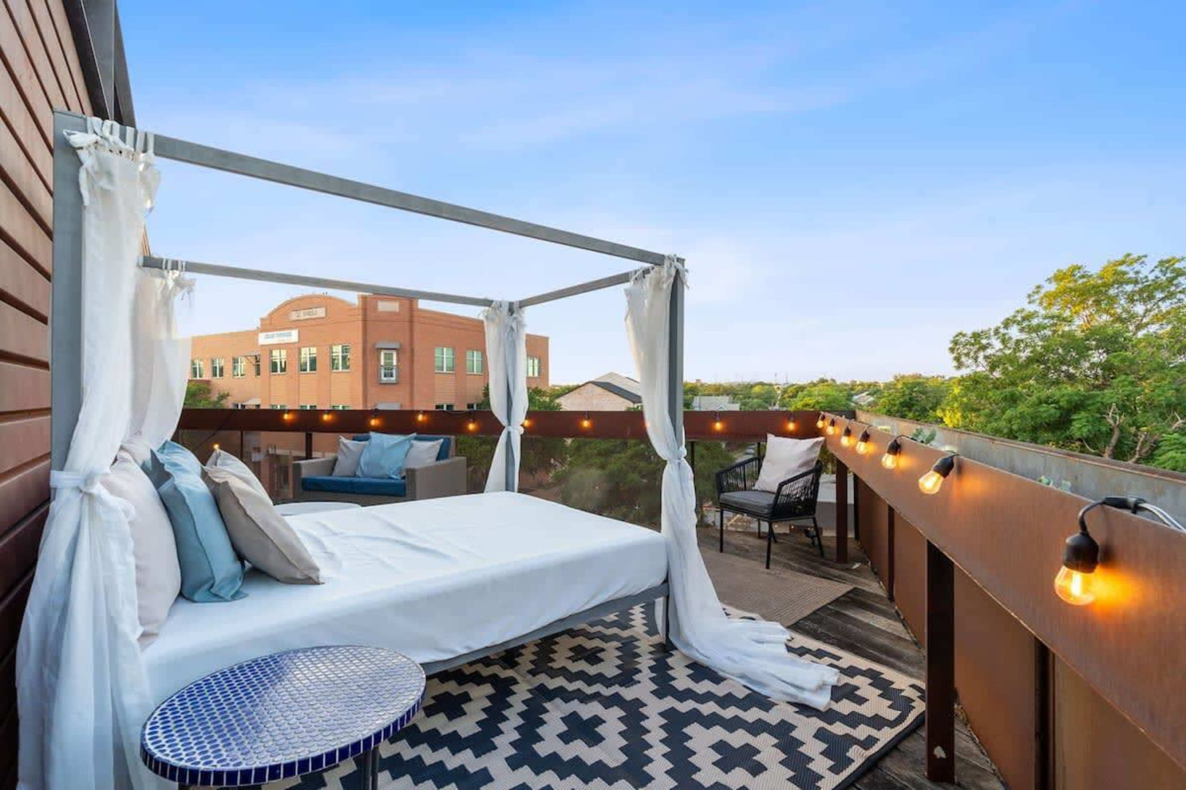 A rooftop terrace featuring a bed with white drapes, surrounded by decorative lights and a patterned rug.