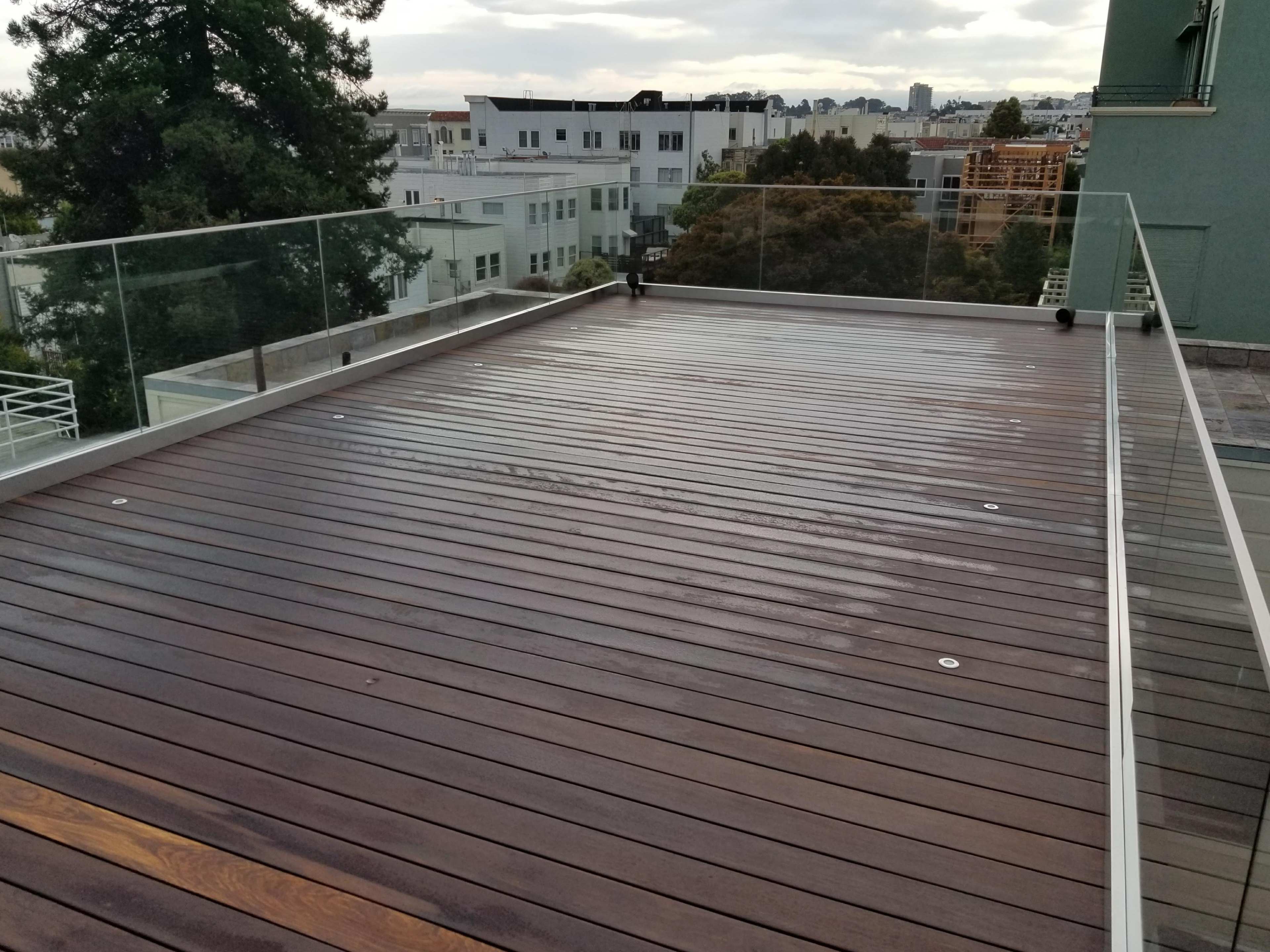 A wooden deck with a glass railing, situated on a rooftop overlooking buildings and trees under a cloudy sky.
