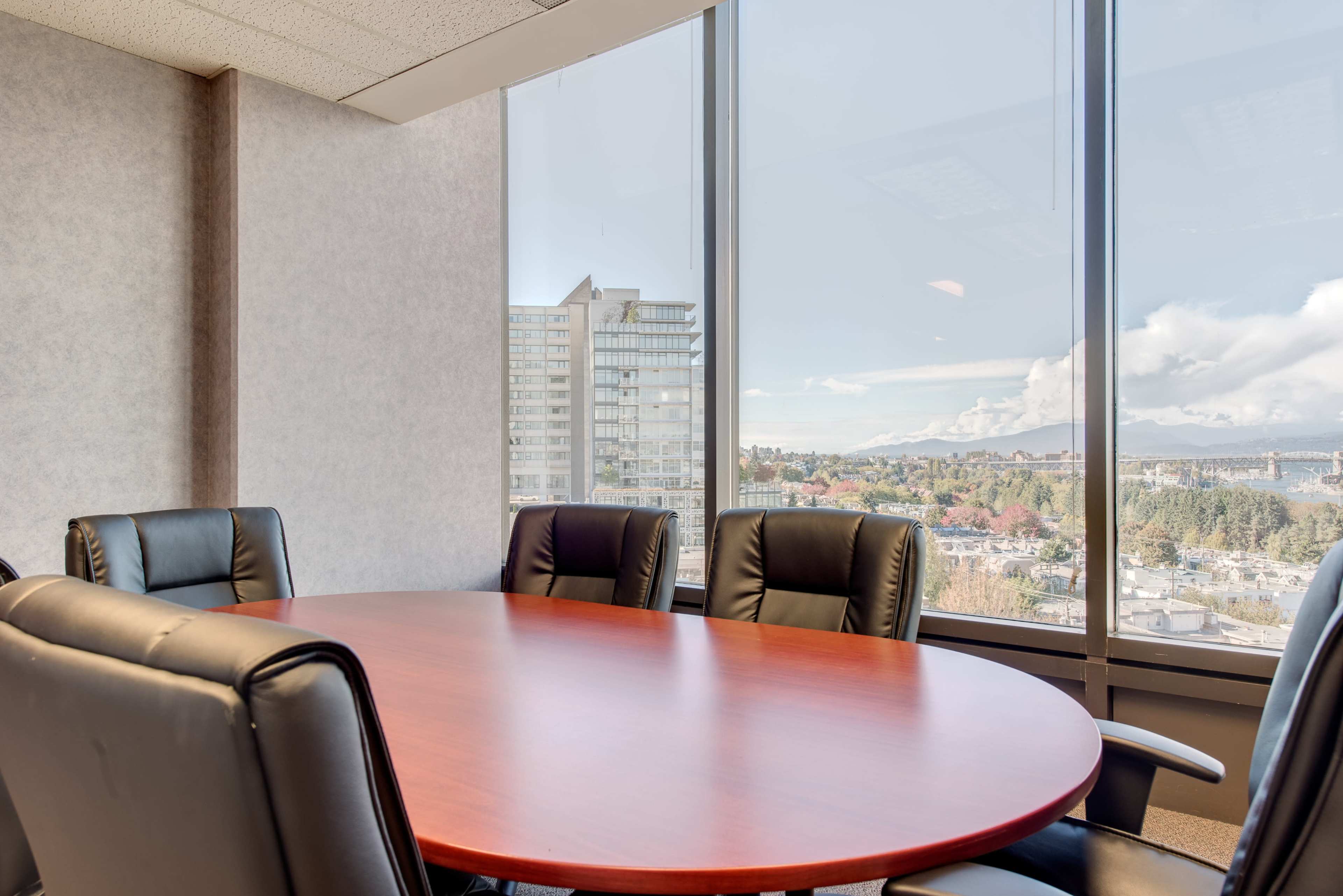 A modern conference room with a round wooden table surrounded by black leather chairs and a large window displaying a cityscape and distant mountains.