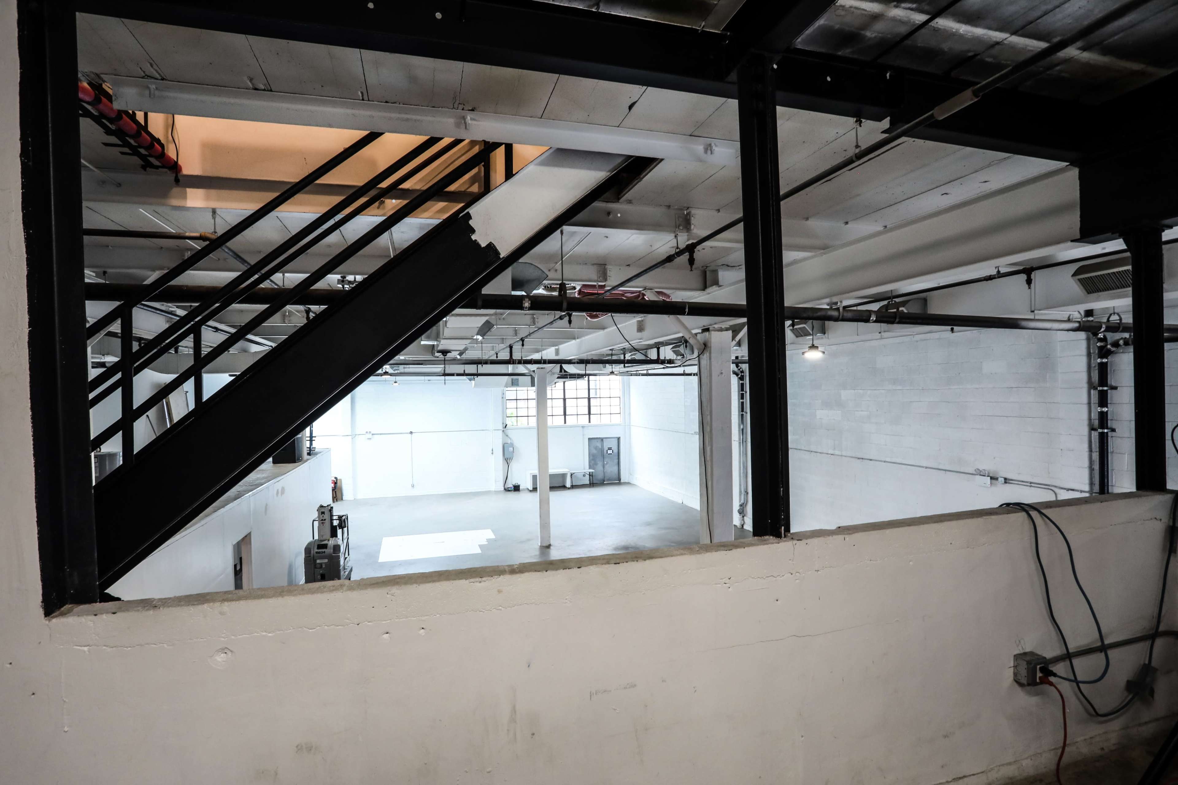 An empty industrial space with large windows, a staircase in the foreground, and white walls.