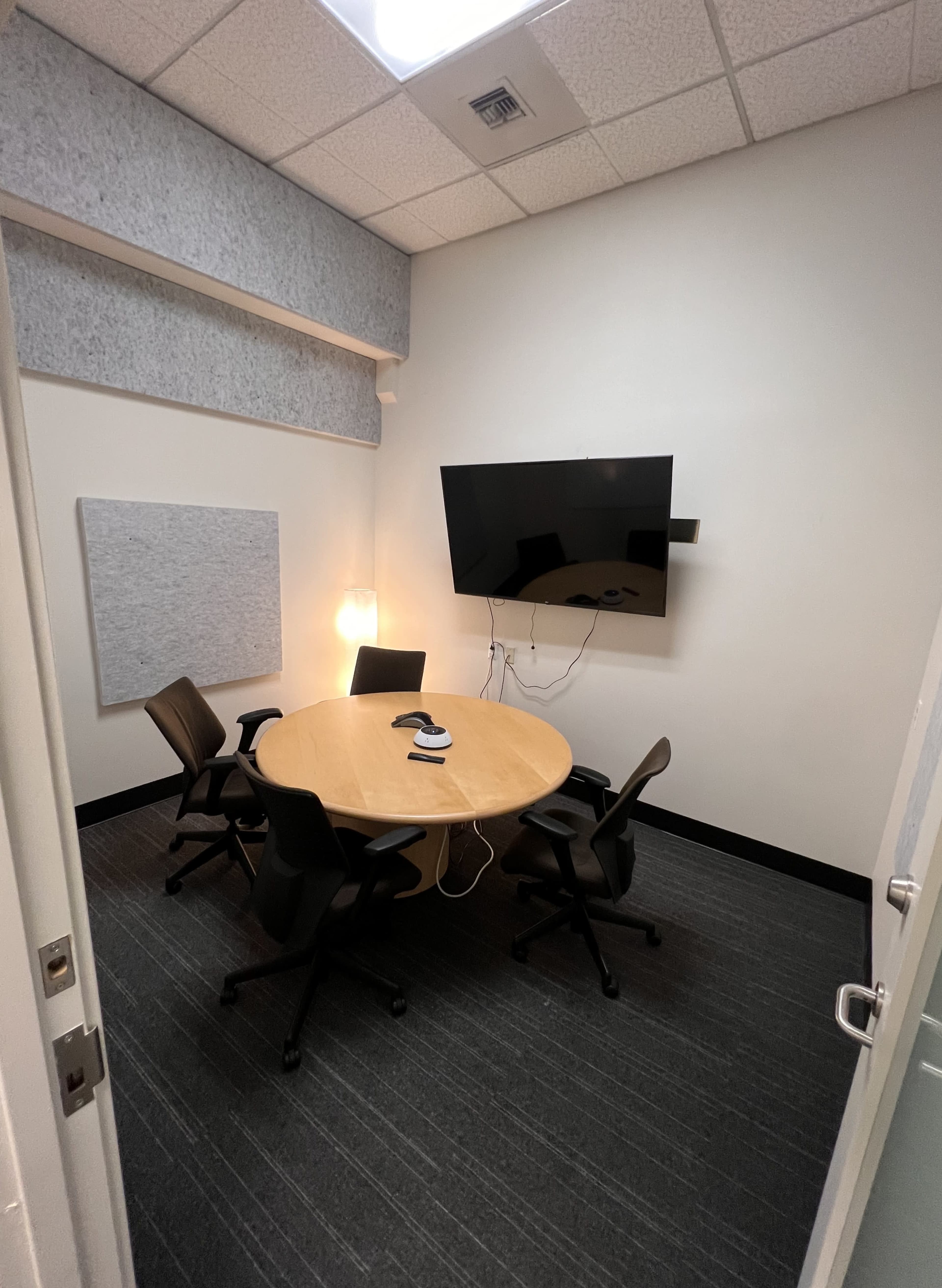 The image shows a small conference room with a round wooden table, four chairs, a wall-mounted television, and a light fixture.