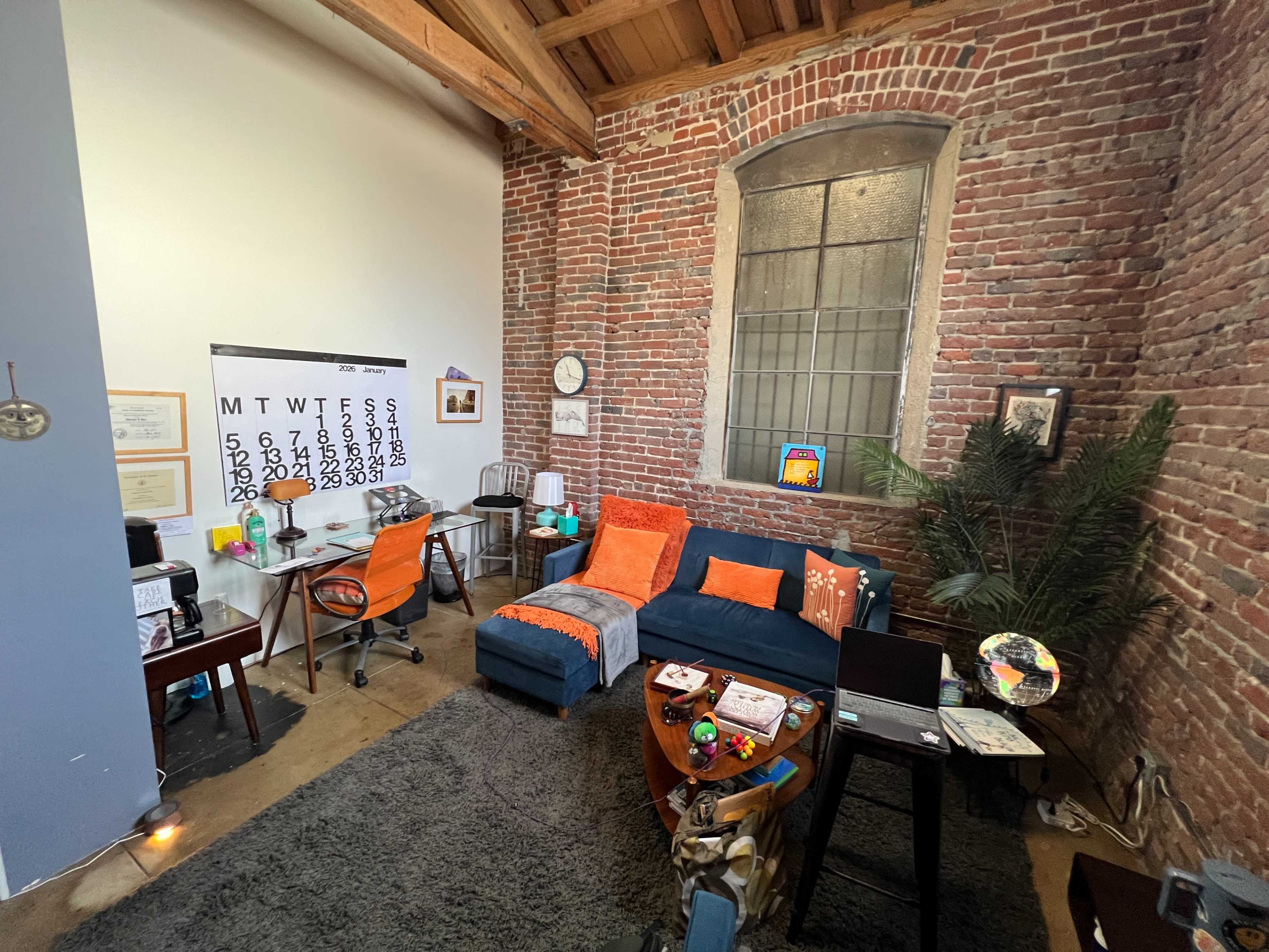 The image shows a cozy workspace featuring a blue sofa with orange throw pillows, a desk with a calendar, and exposed brick walls in a well-lit room.