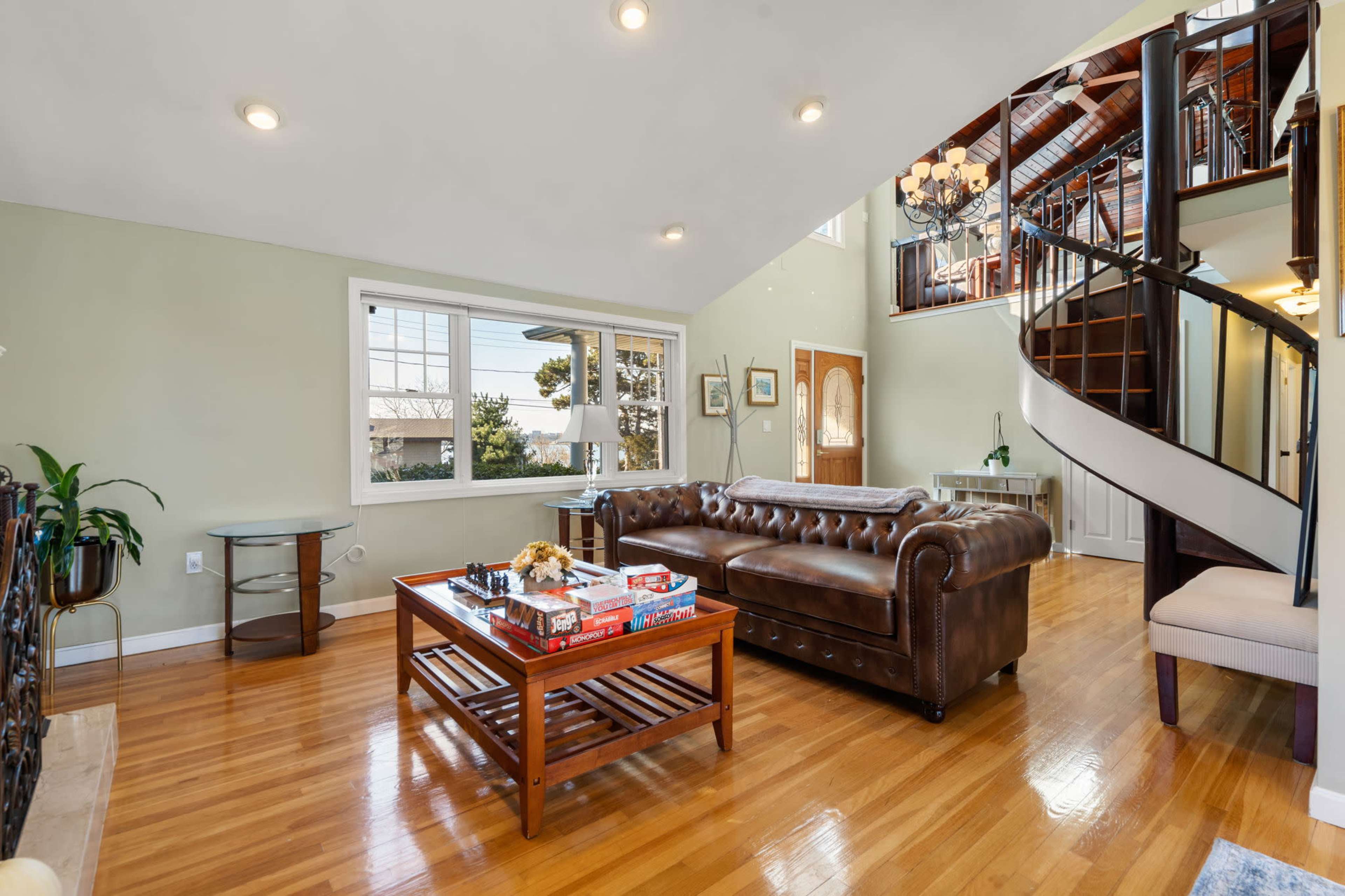 A spacious living room features a brown leather sofa, a wooden coffee table with board games, and a winding staircase leading to an upper level.