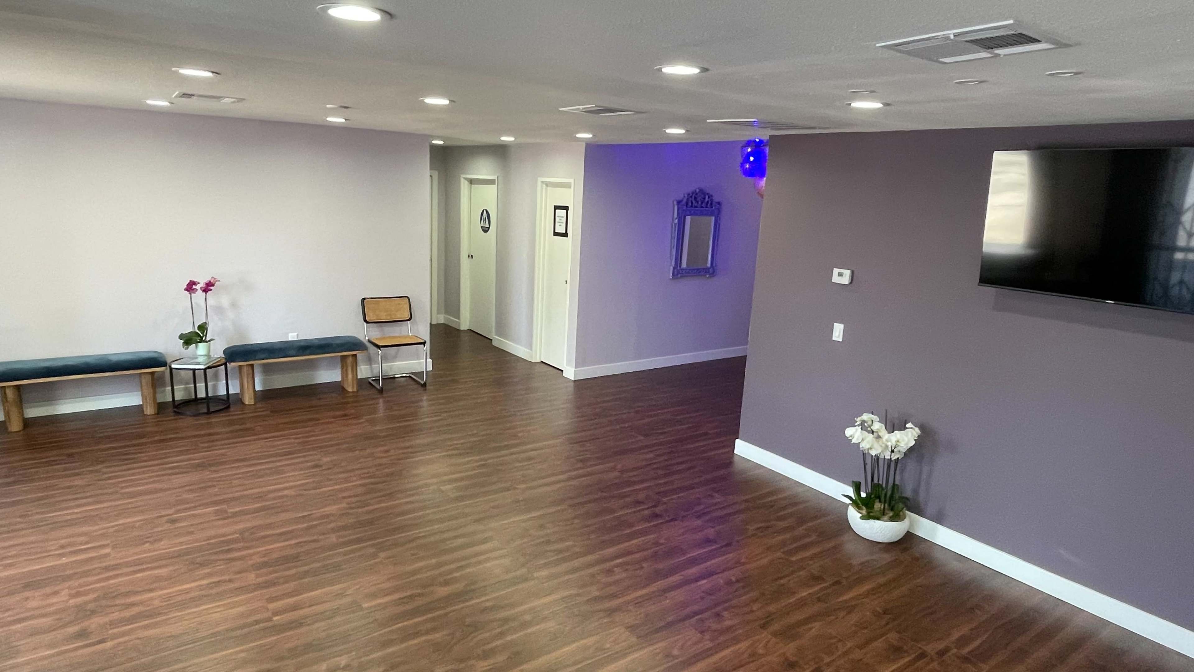 A spacious, well-lit interior with wooden flooring, featuring two benches, a television on the wall, and a hallway leading to closed doors.