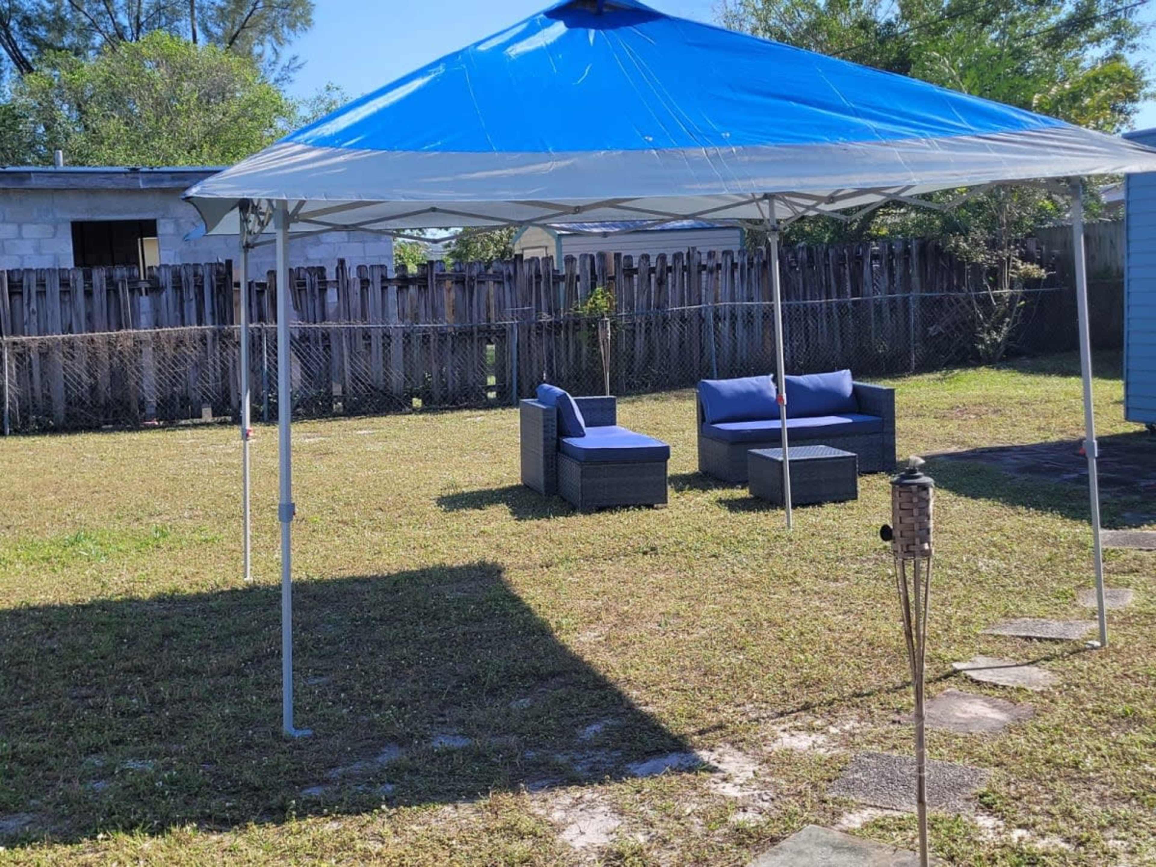 A blue and white canopy covers two outdoor couches and a table on a grassy area surrounded by a fence and trees.