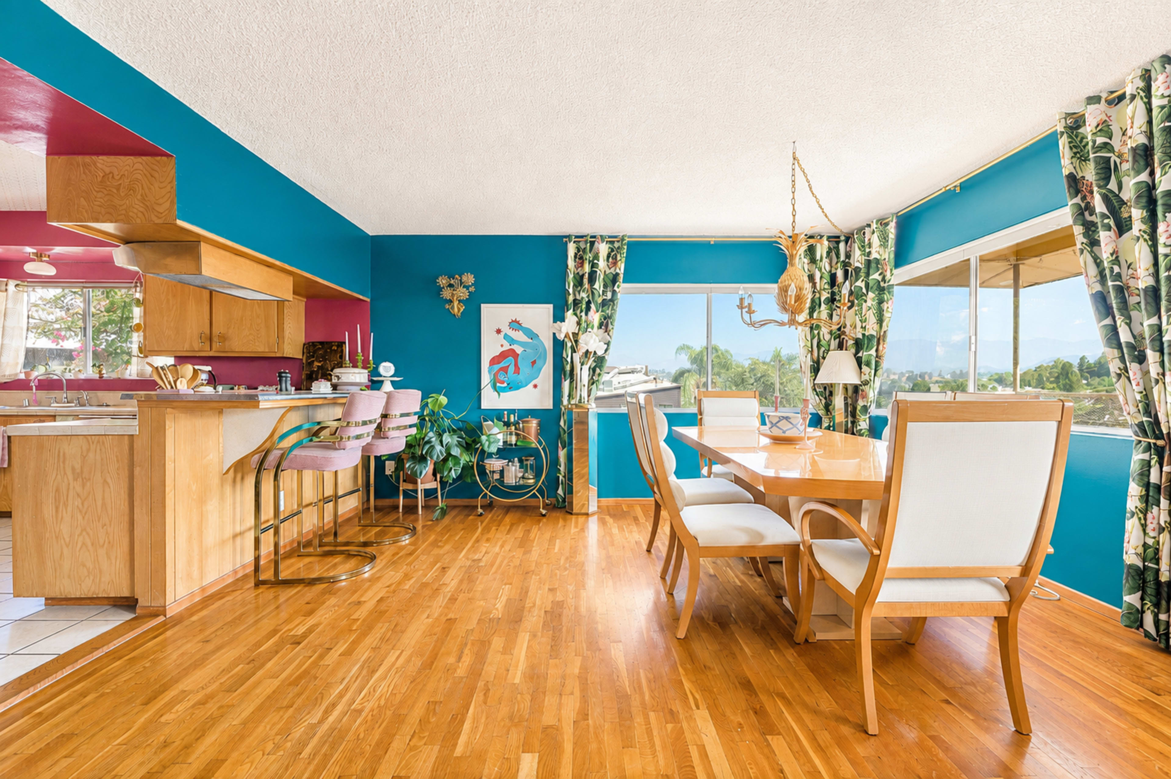 The image shows a bright dining area with a wooden table and chairs, adjacent to a kitchen bar with stools, surrounded by large windows and colorful decor.