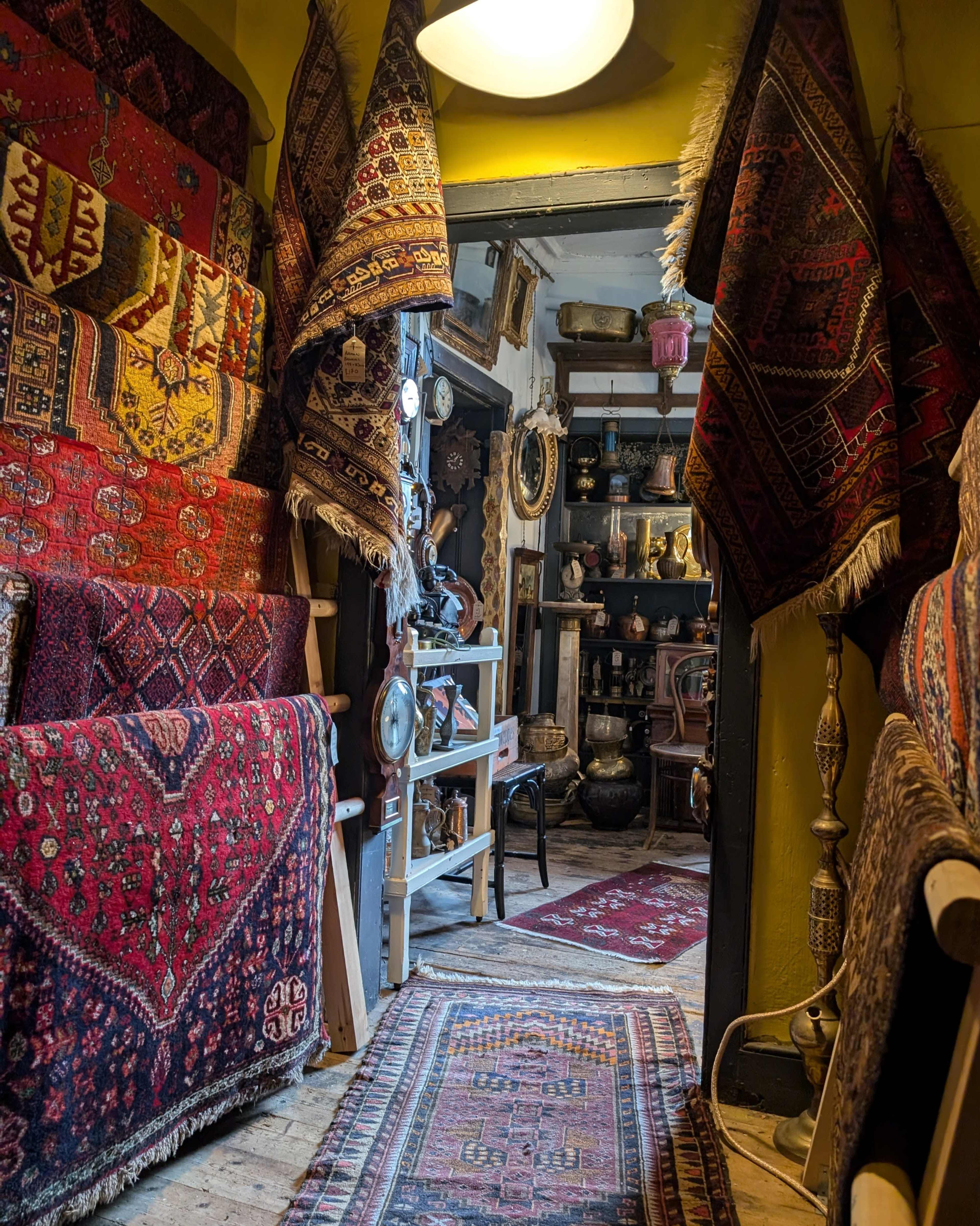 The image shows a narrow passageway leading into a room filled with colorful rugs and various decorative items.
