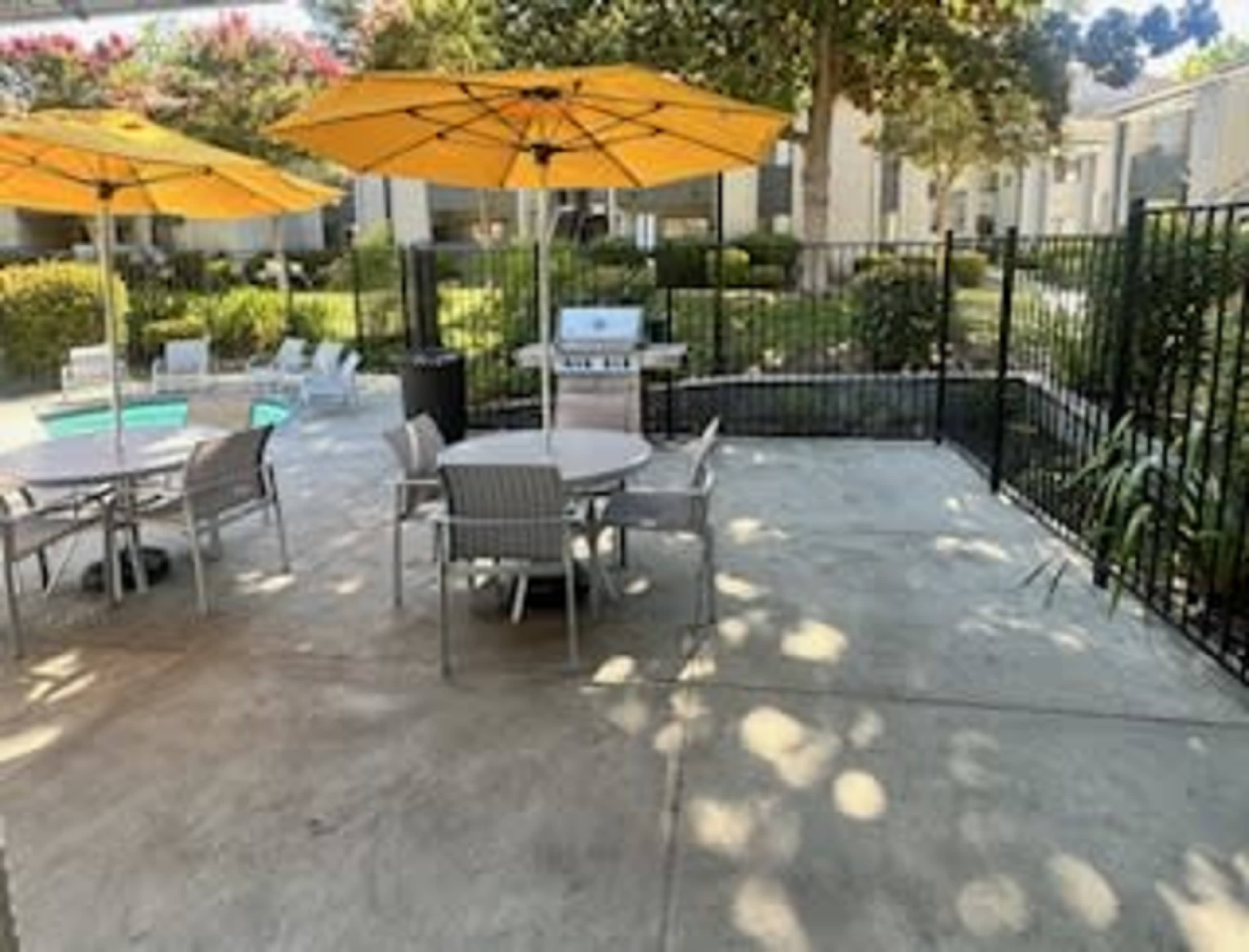 A patio area with round tables and chairs under yellow umbrellas, near a barbecue grill and surrounded by greenery and a swimming pool.