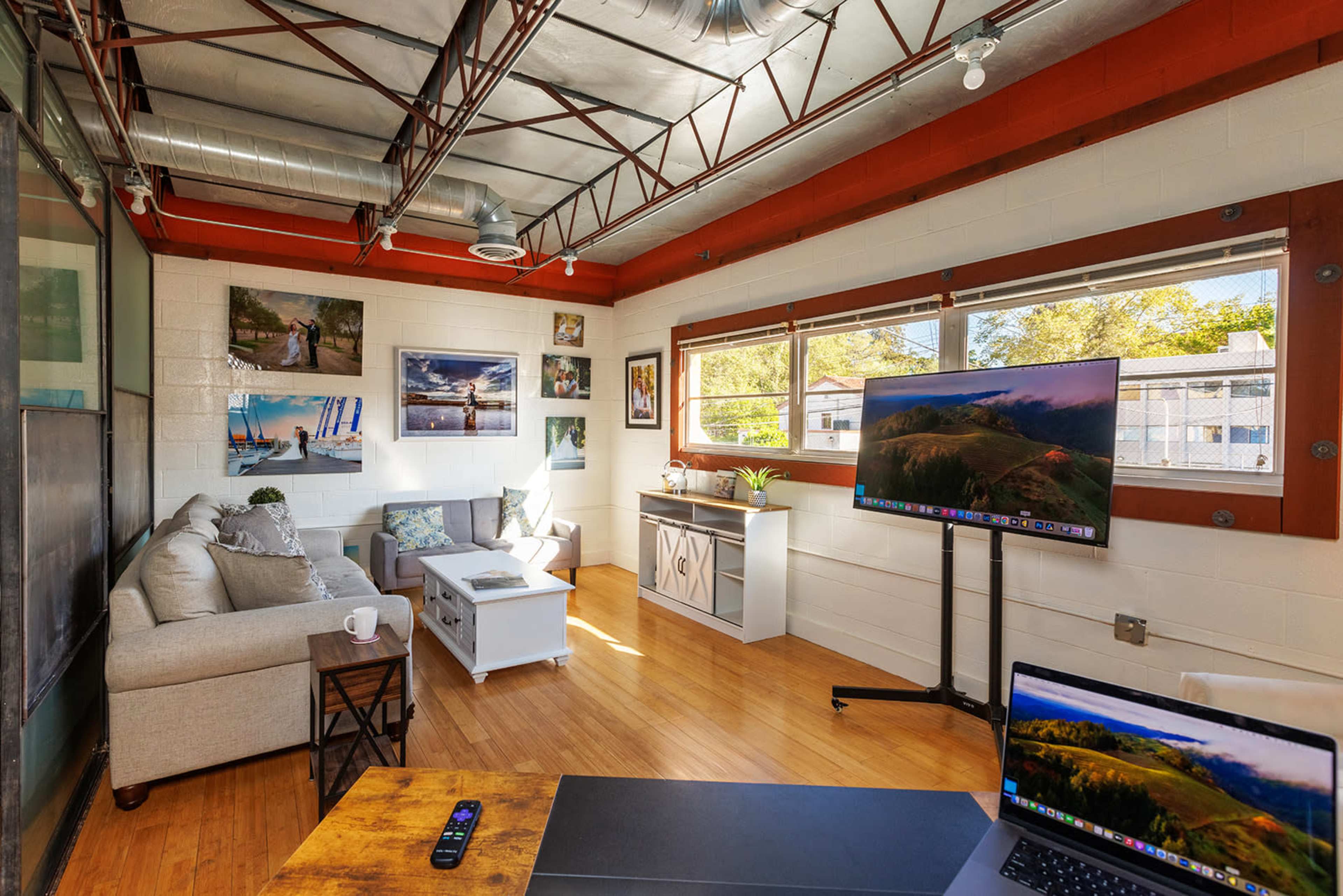 A modern office space featuring a sofa, a coffee table, a large monitor on a stand, and several framed photographs on the walls.