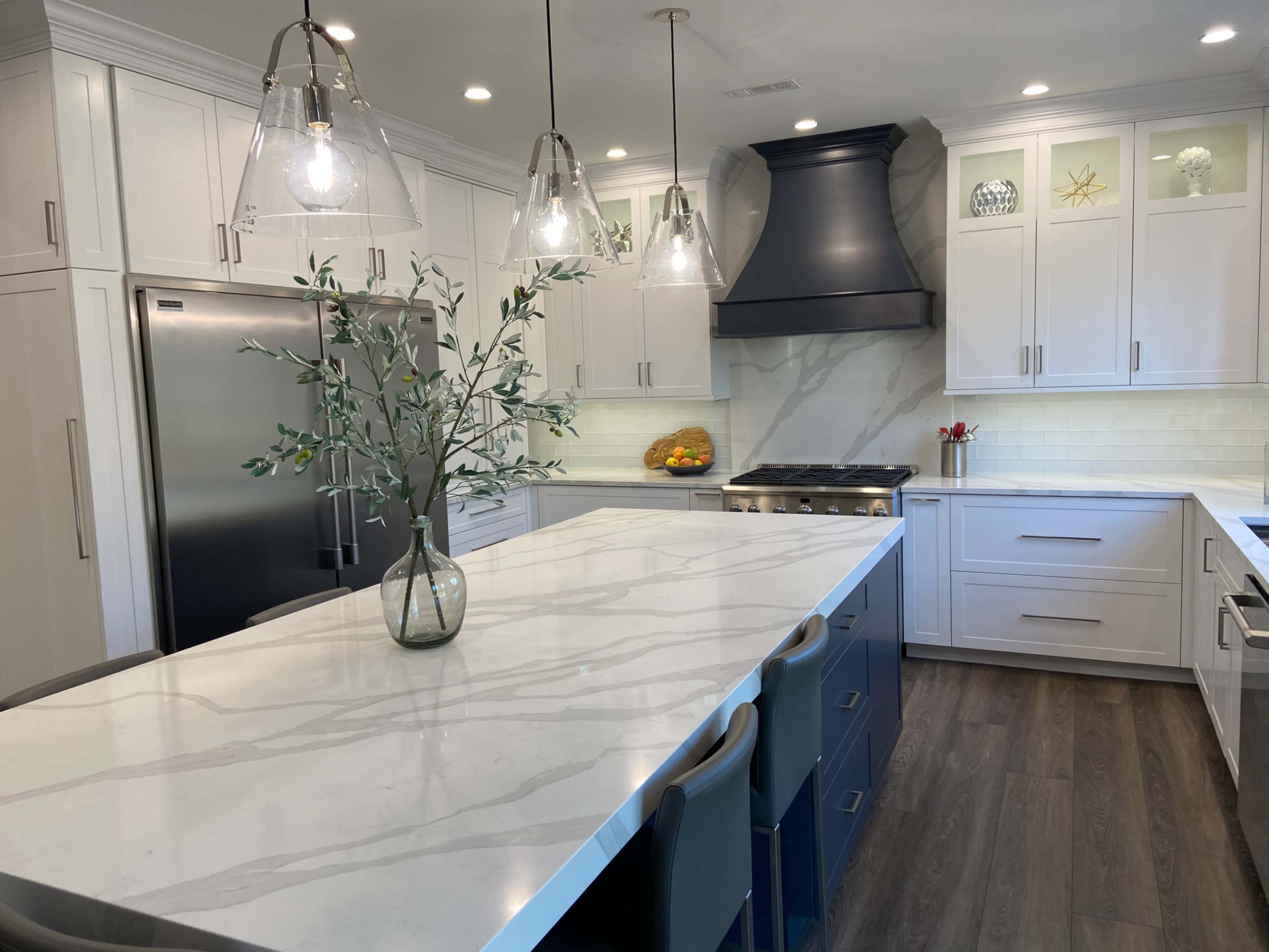 A modern kitchen features a large island with a marble countertop, white cabinetry, stainless steel appliances, and pendant lighting.