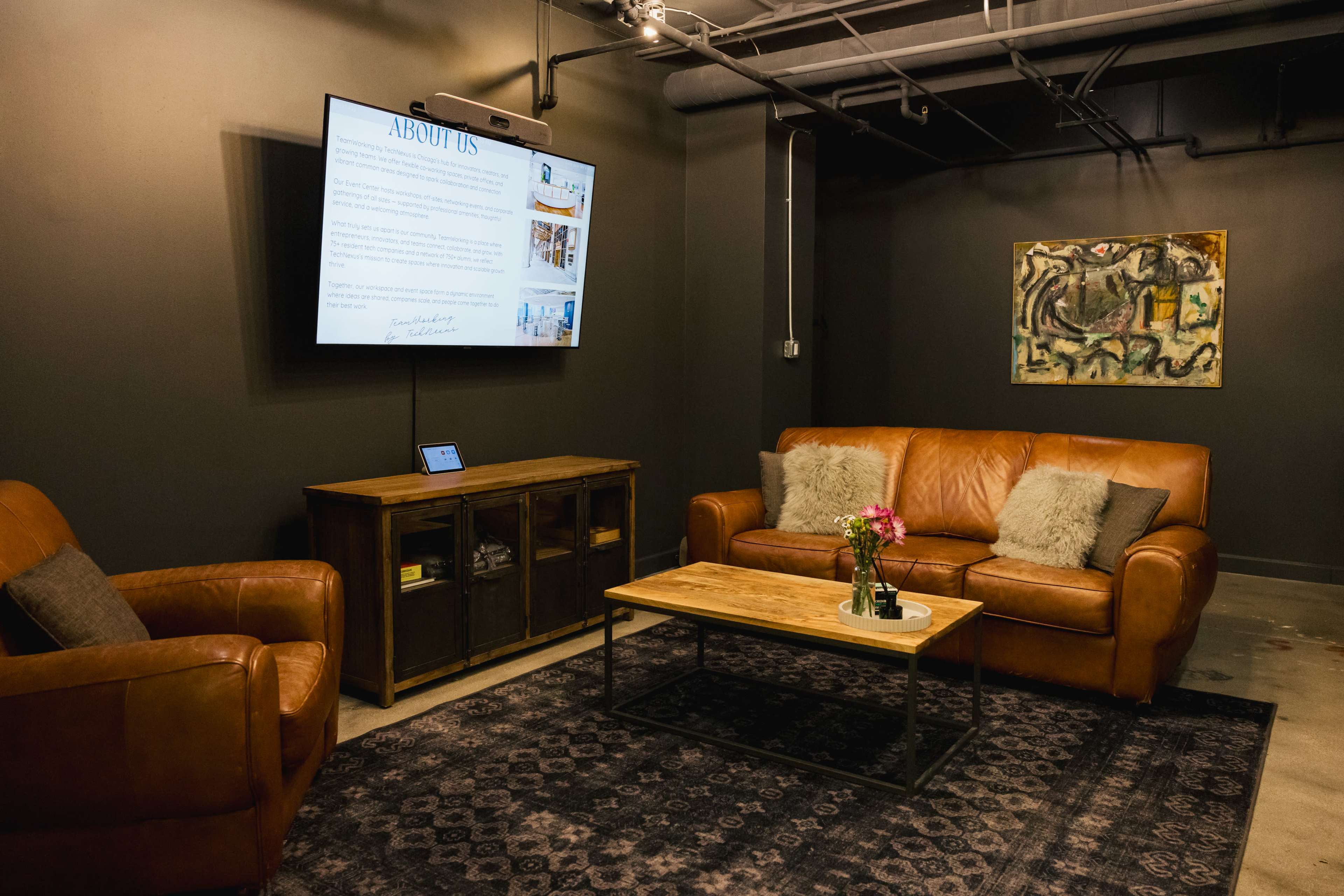 The image depicts a modern lounge area with two brown leather sofas, a wooden coffee table, a dark rug, and a wall-mounted screen displaying information.