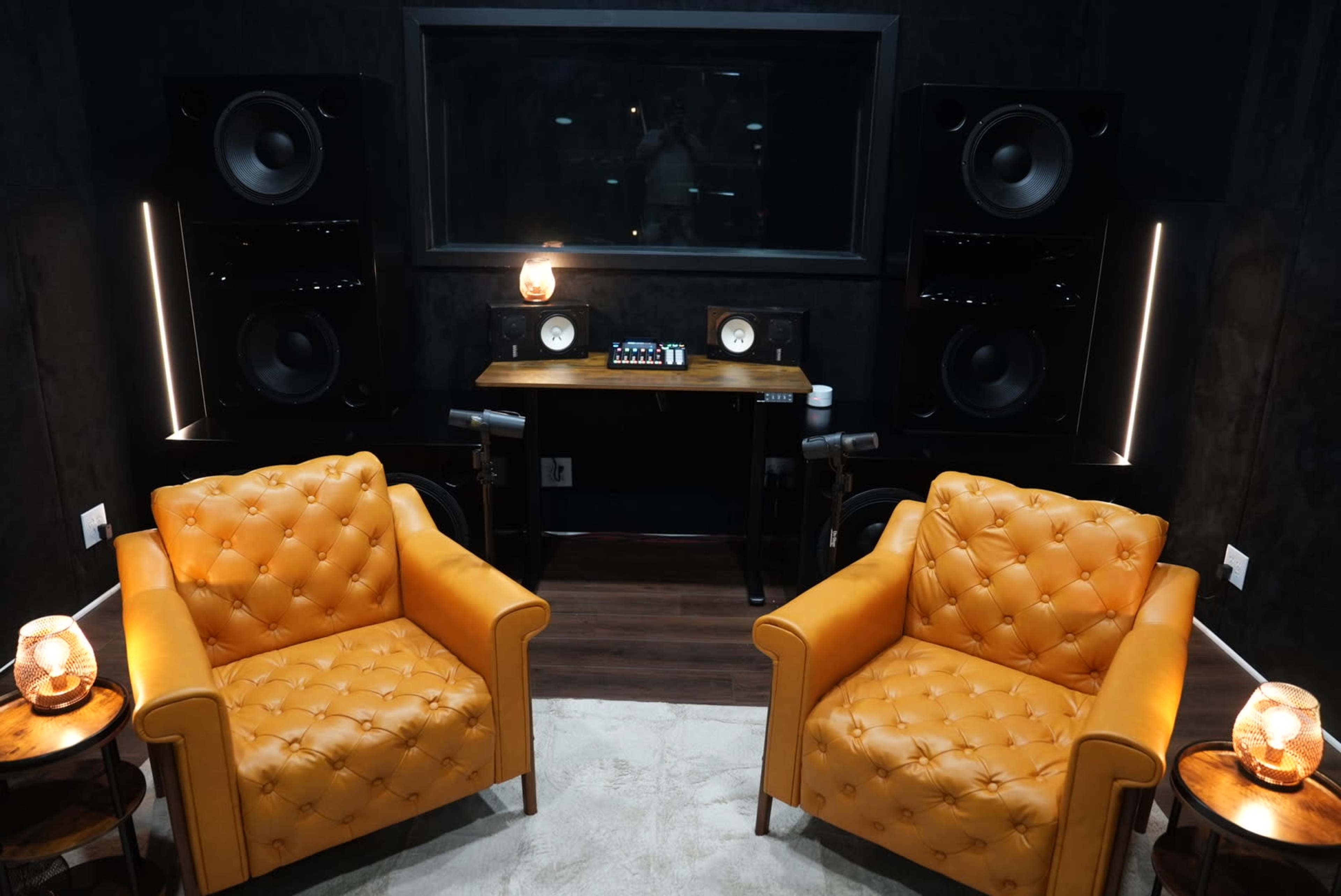 The image shows a cozy recording studio featuring two orange tufted armchairs, a wooden work desk with audio equipment, and large speakers on either side of a glass panel.