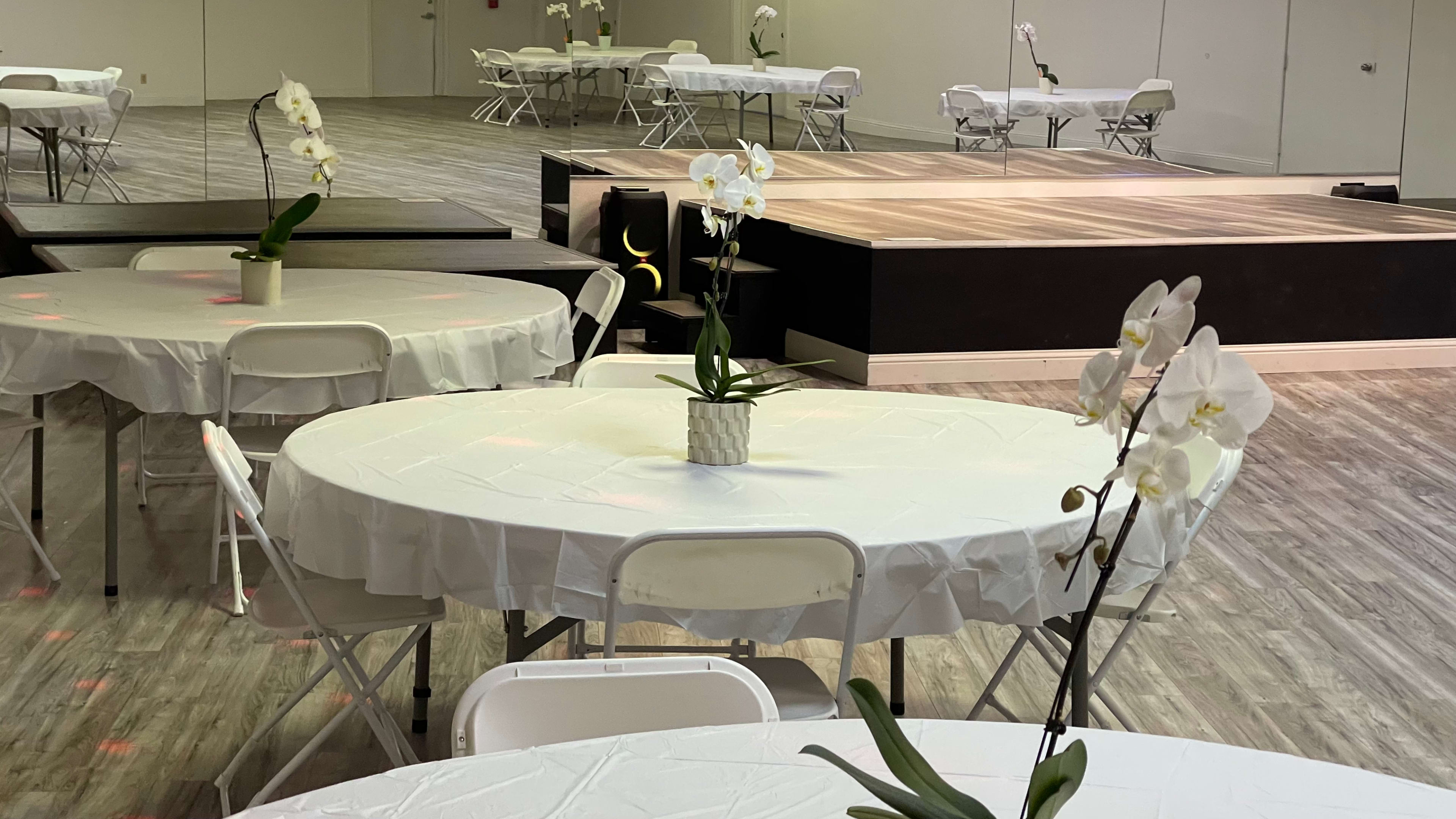 A spacious event room with white tablecloth-covered round tables, each featuring a potted orchid, and a small stage at one end.