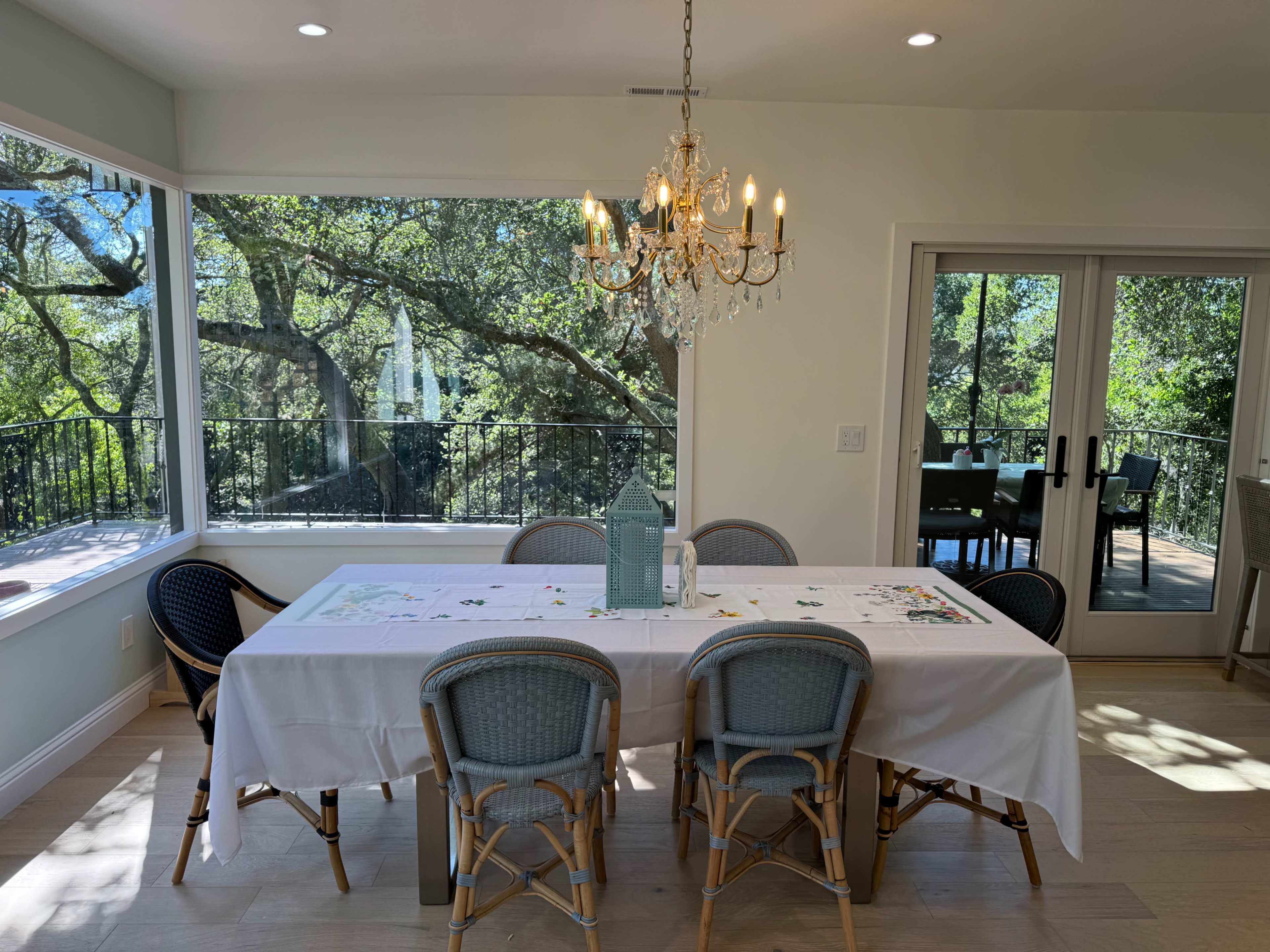 A dining area features a long white table surrounded by eight chairs, with a chandelier overhead and large windows showcasing a view of trees outside.
