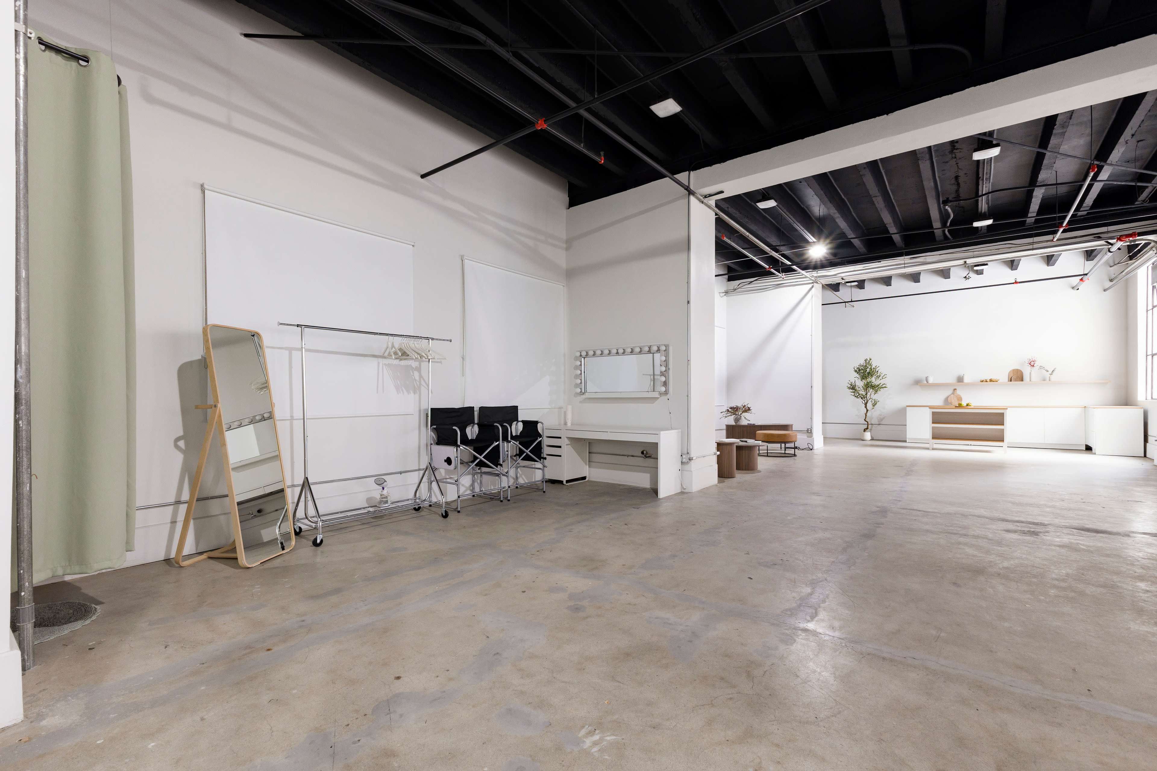 The image shows an empty studio space featuring a clothing rack, a large mirror, a white table against the wall, and minimal furnishings, all set against a stark, industrial backdrop.