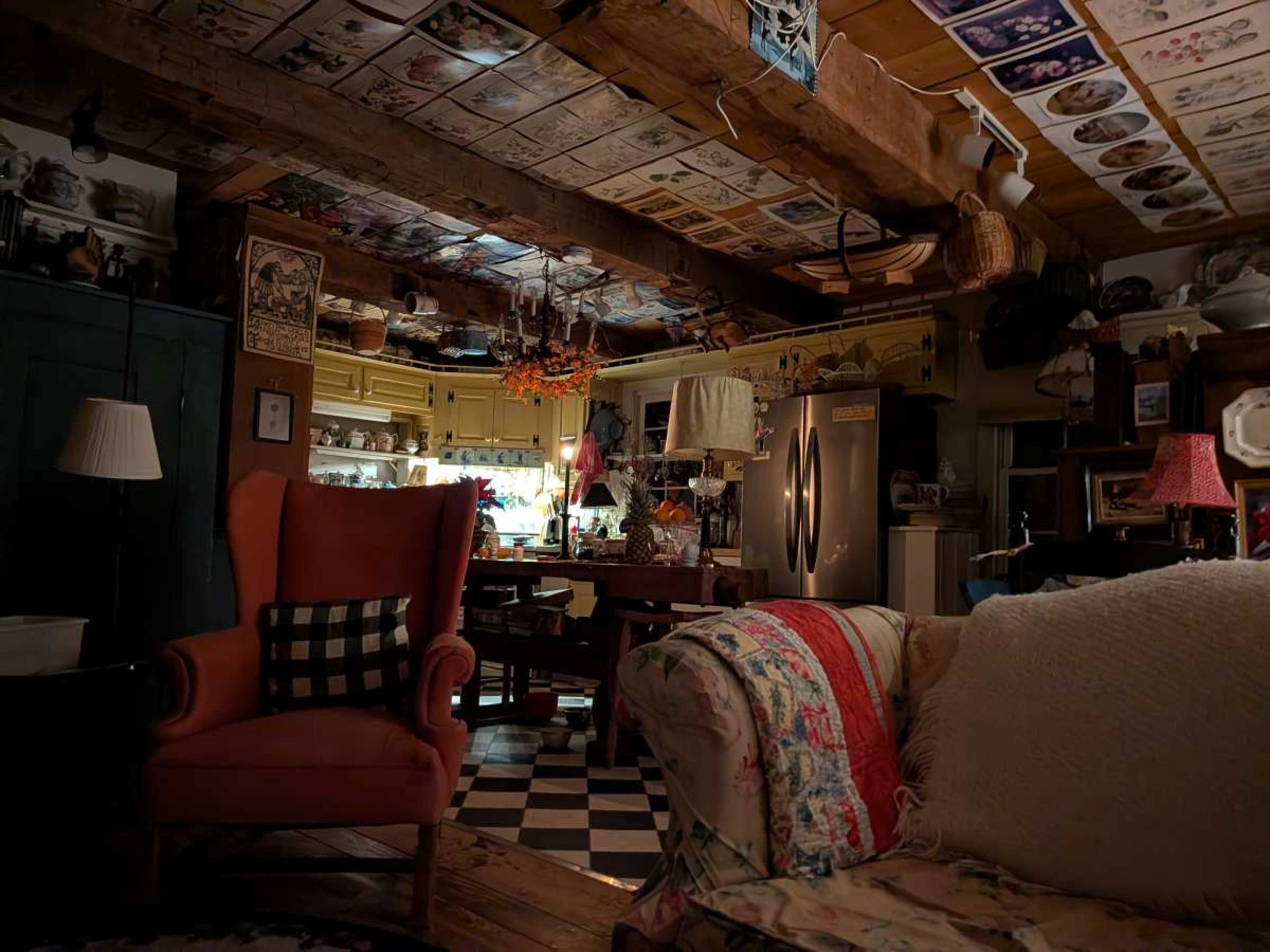The image shows a cozy kitchen with wooden beams, vintage decor on the ceiling, and a mix of furniture, including a pink armchair and a colorful couch.