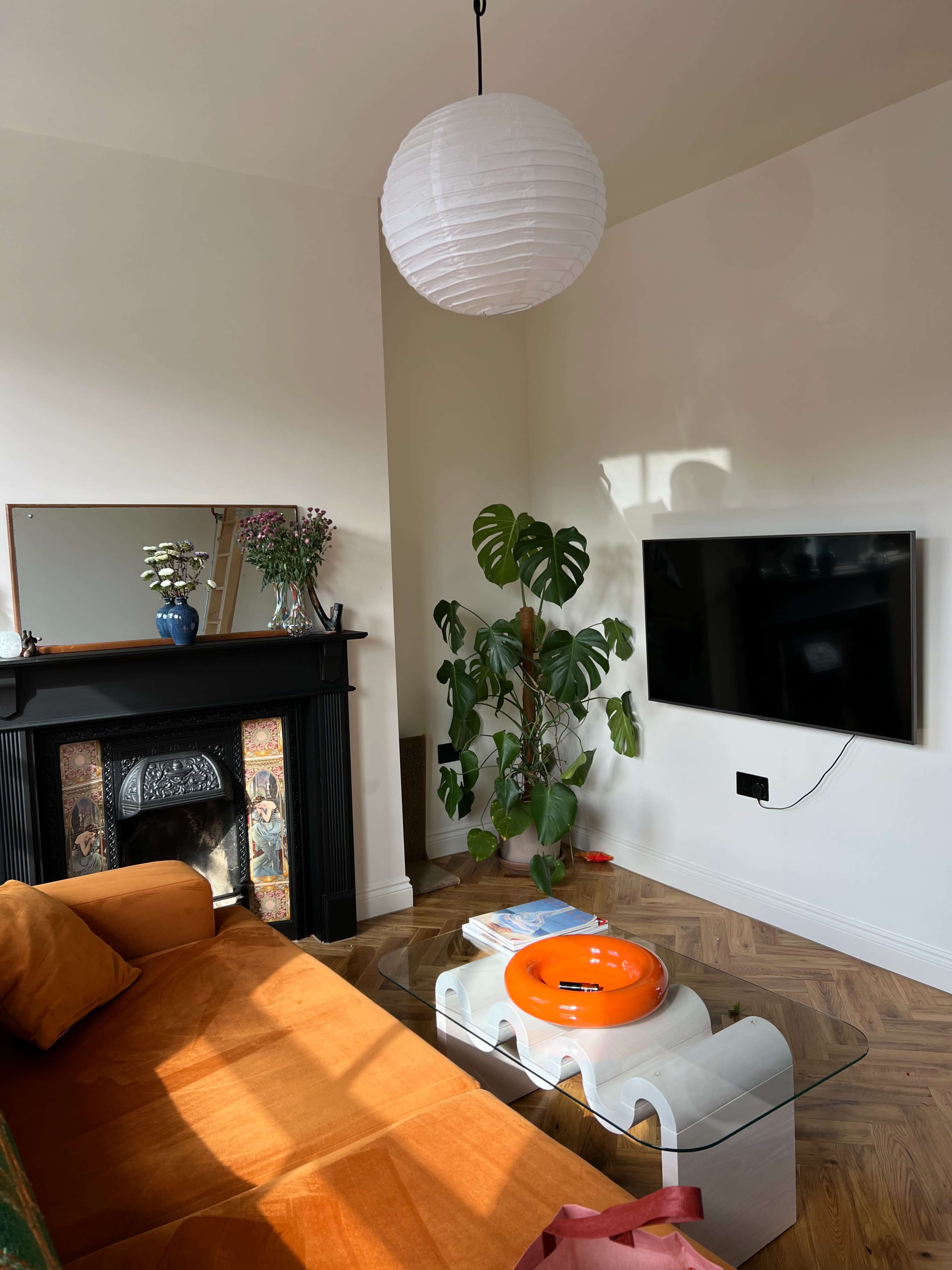 A modern living room features a bright orange sofa, a glass coffee table with an orange bowl, a television mounted on the wall, and a potted plant in the corner, all illuminated by natural light.