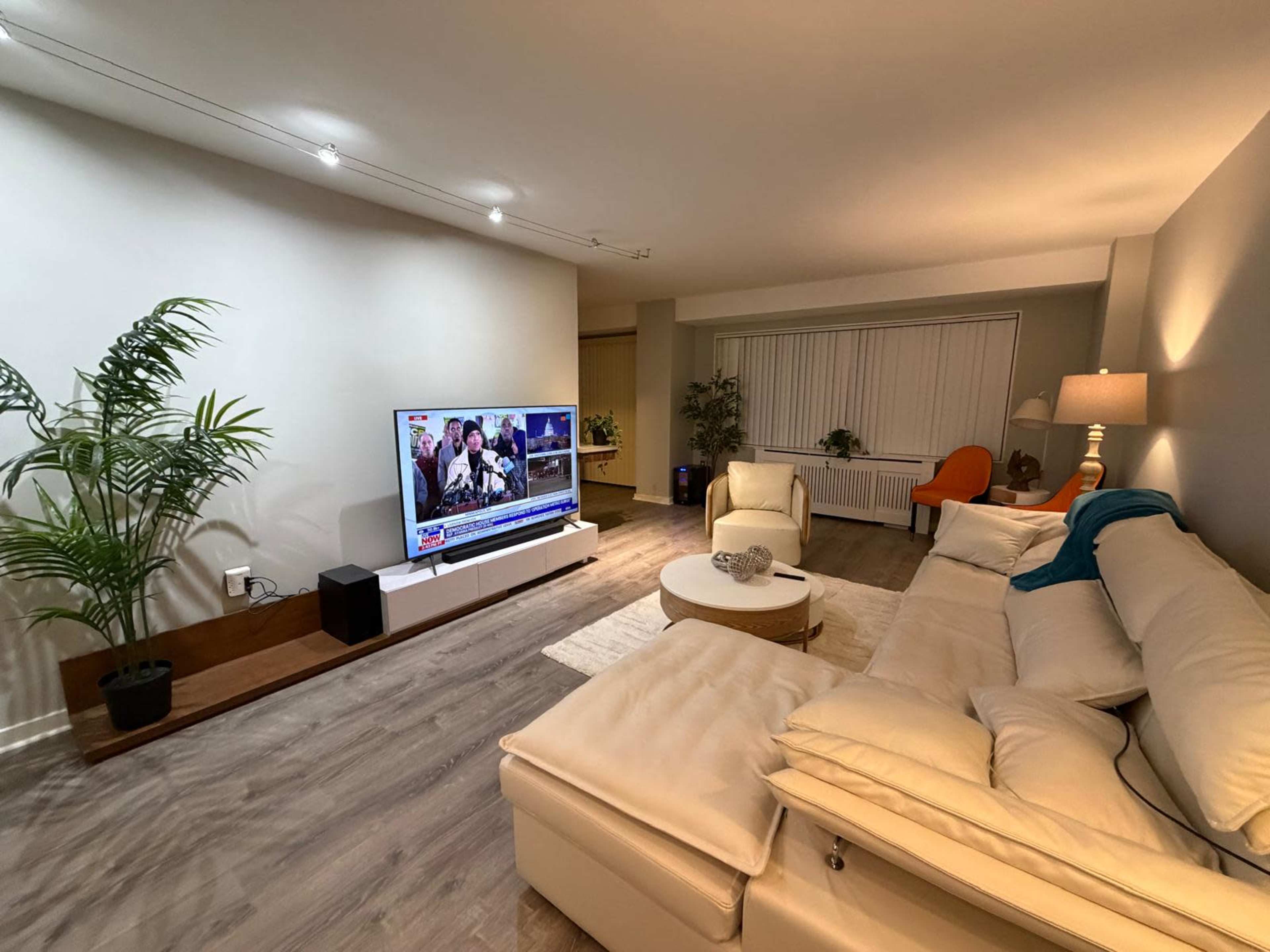 A modern living room features a white sectional sofa, a round coffee table, a flat-screen TV mounted on the wall, and a large window with closed blinds.