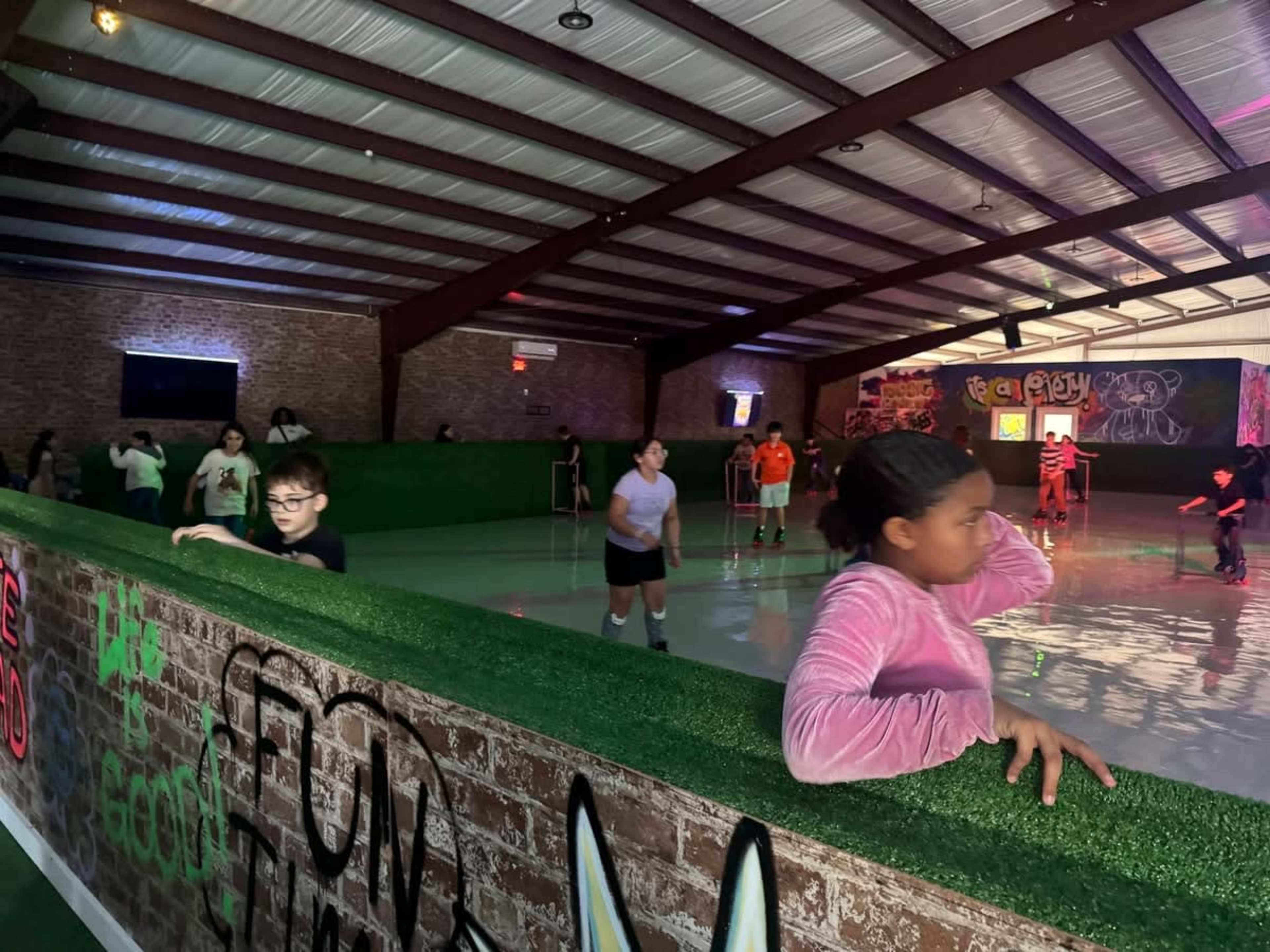 A group of children skate on a polished rink inside a recreational facility decorated with graffiti.