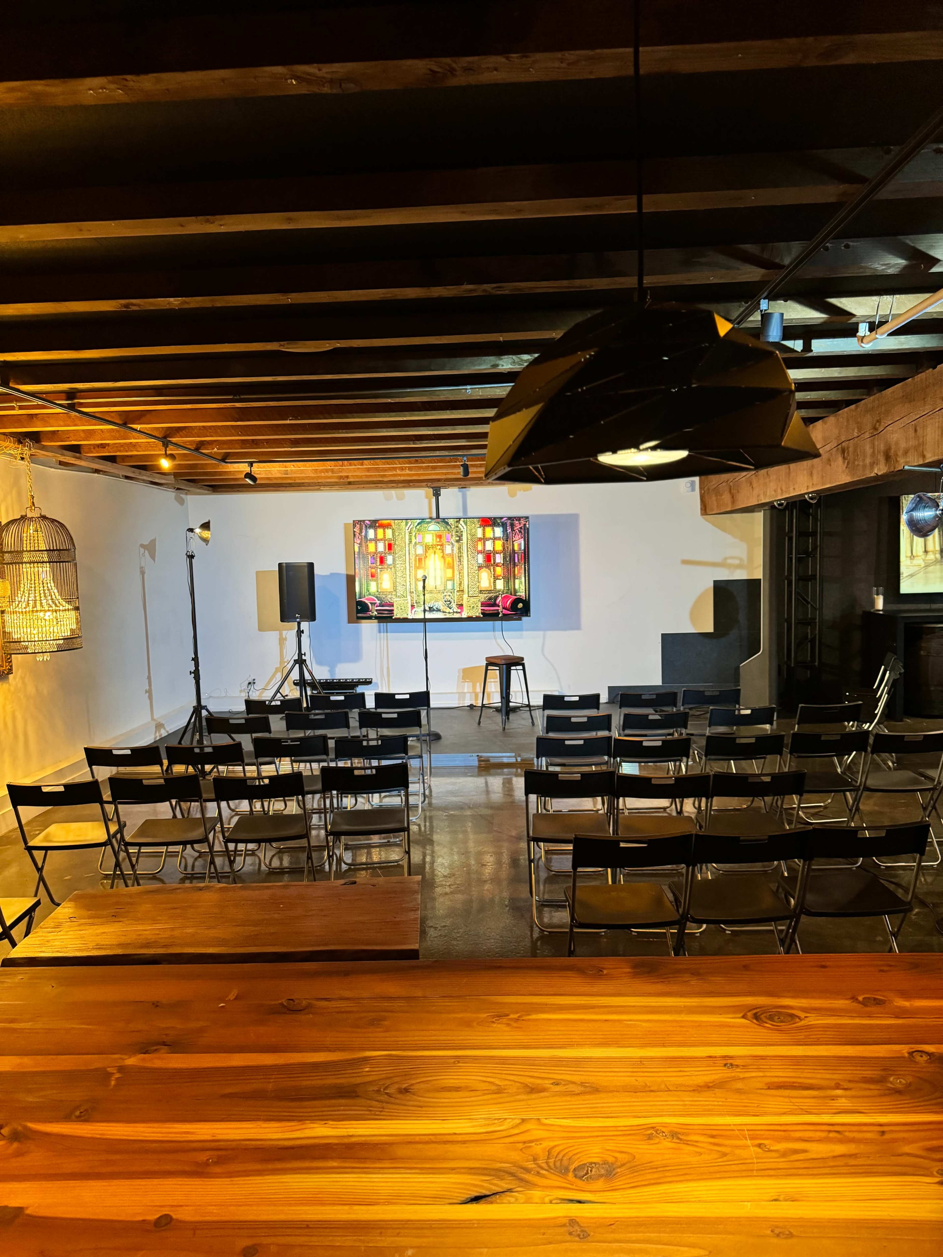A spacious interior setting with rows of black chairs facing a stage adorned with a colorful stained glass panel, surrounded by wooden beams and a long wooden table in the foreground.