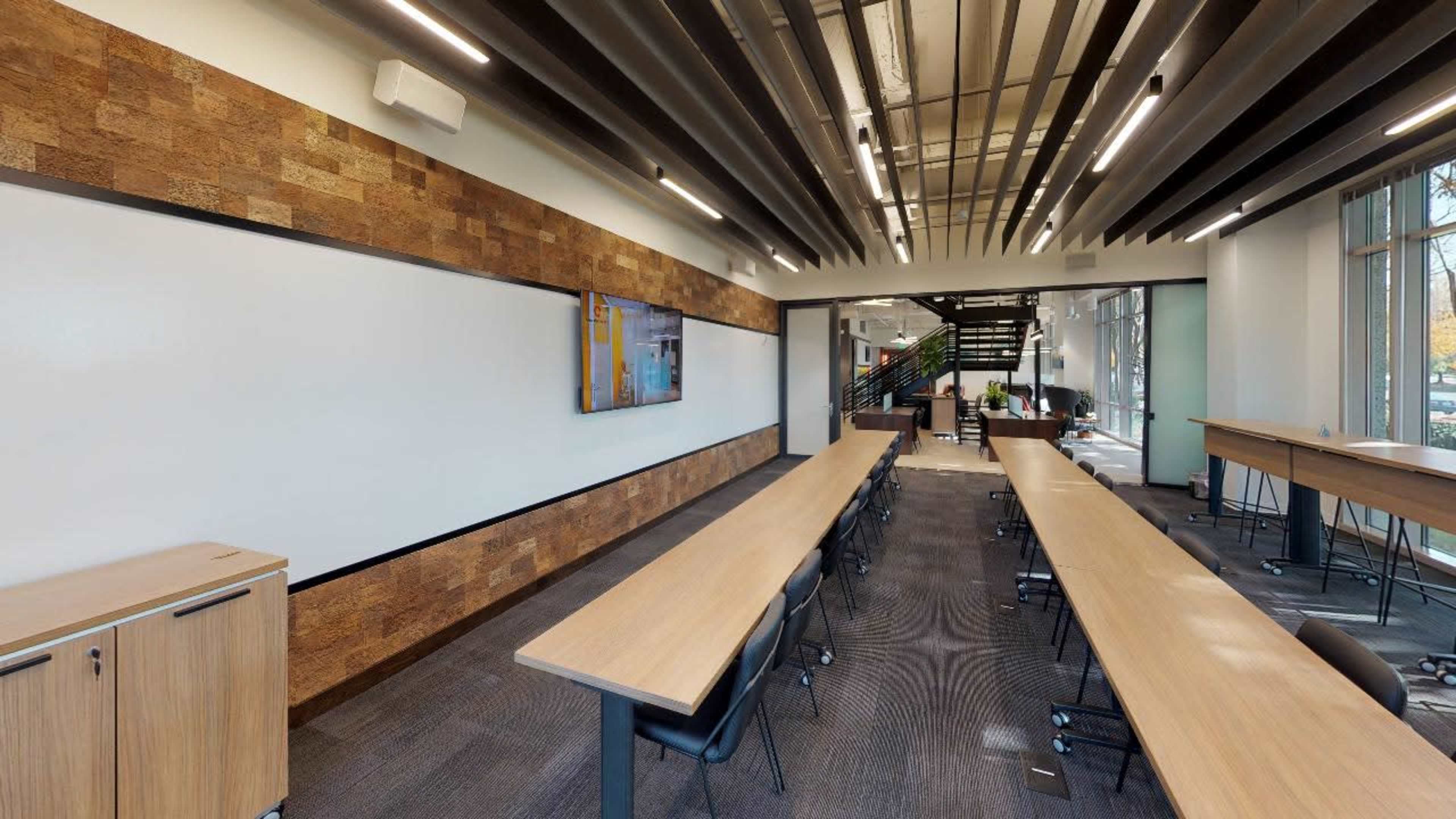 A modern conference room with long wooden tables, a large wall-mounted screen, and large windows allowing natural light.