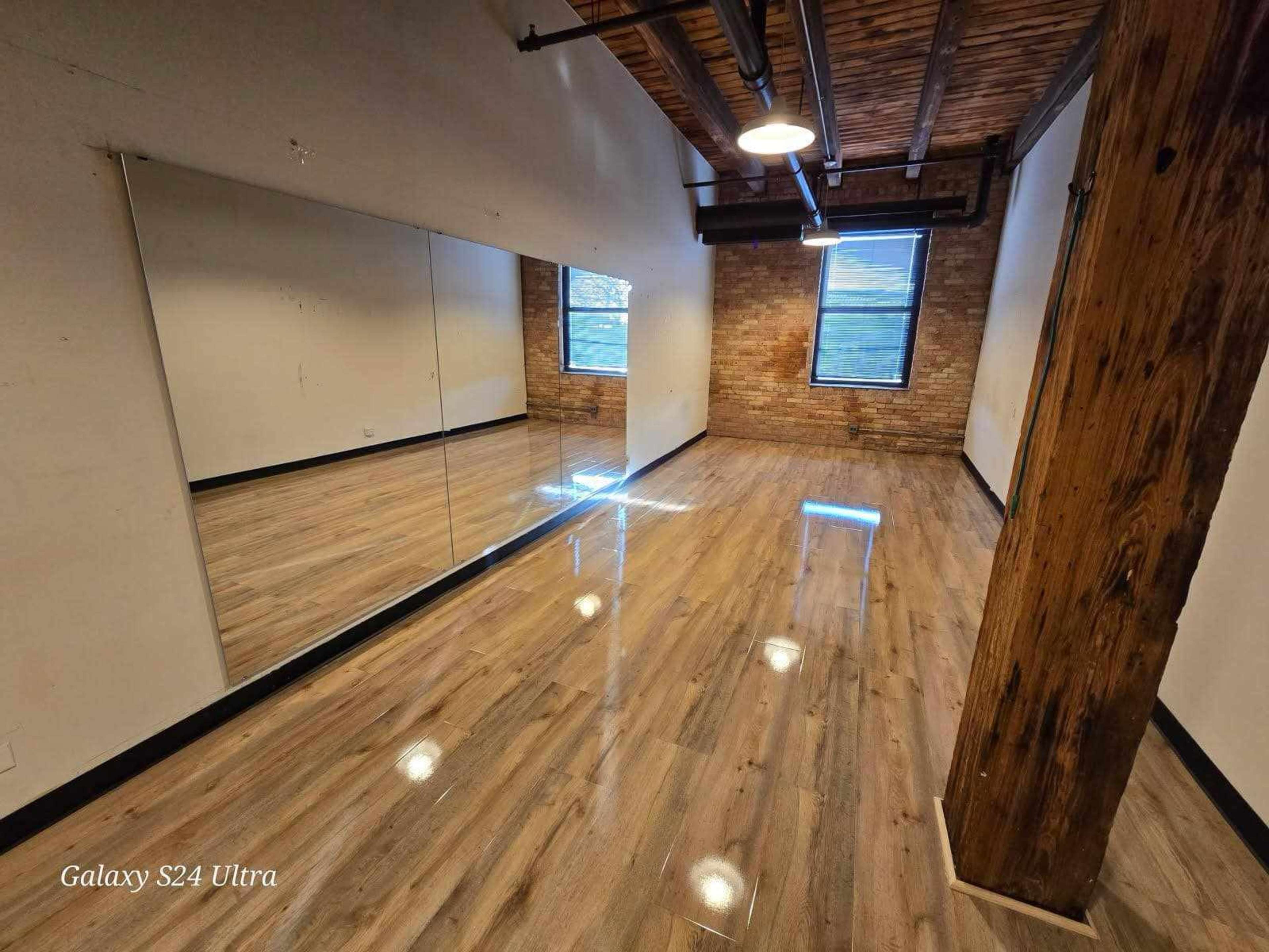 The image shows a spacious room featuring a large mirror on one wall, hardwood flooring, and exposed brick along another wall, with a window allowing natural light.