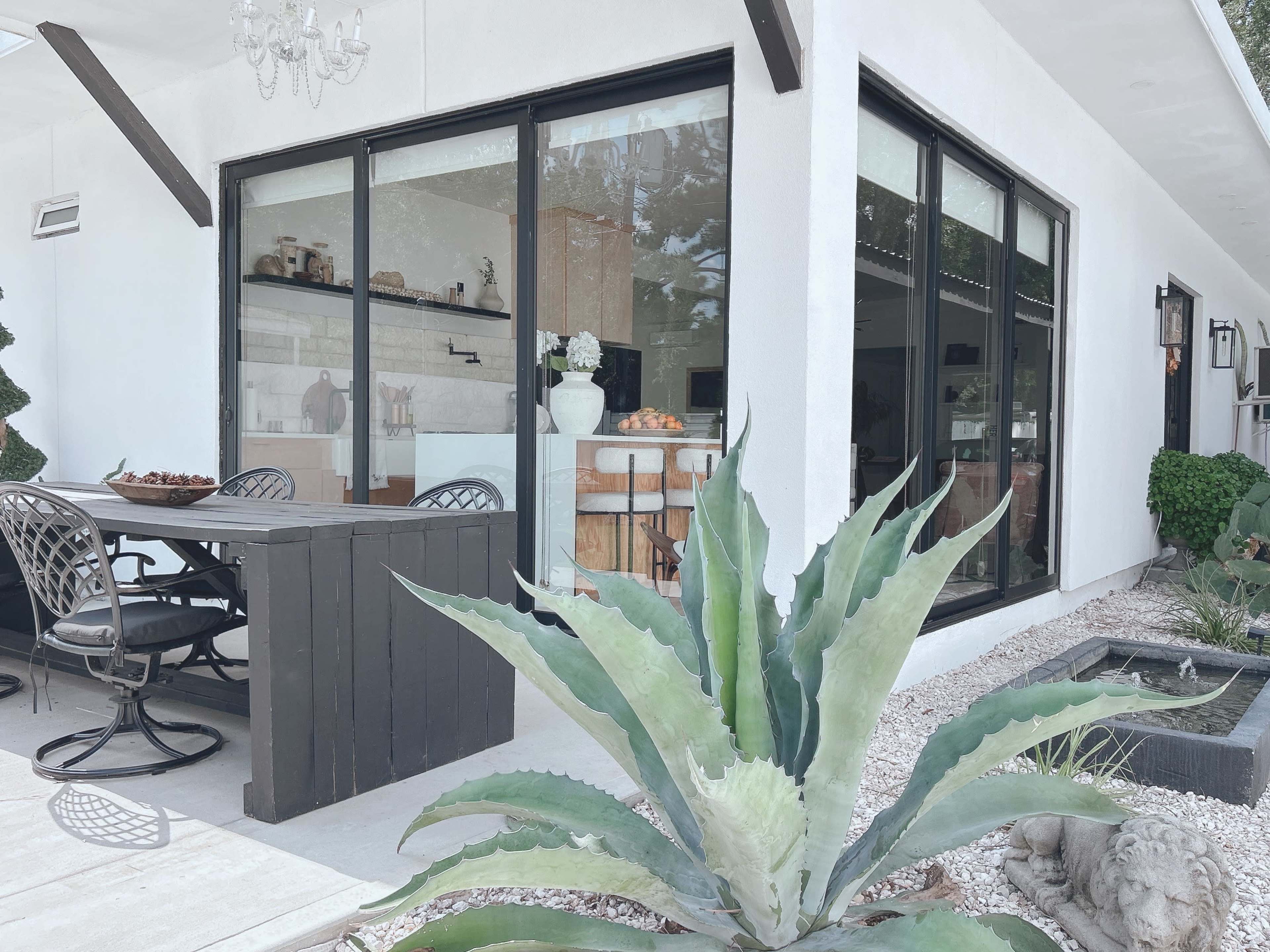 A modern outdoor dining area with a table and chairs, surrounded by potted plants and a small water feature, is adjacent to a glass-enclosed room.