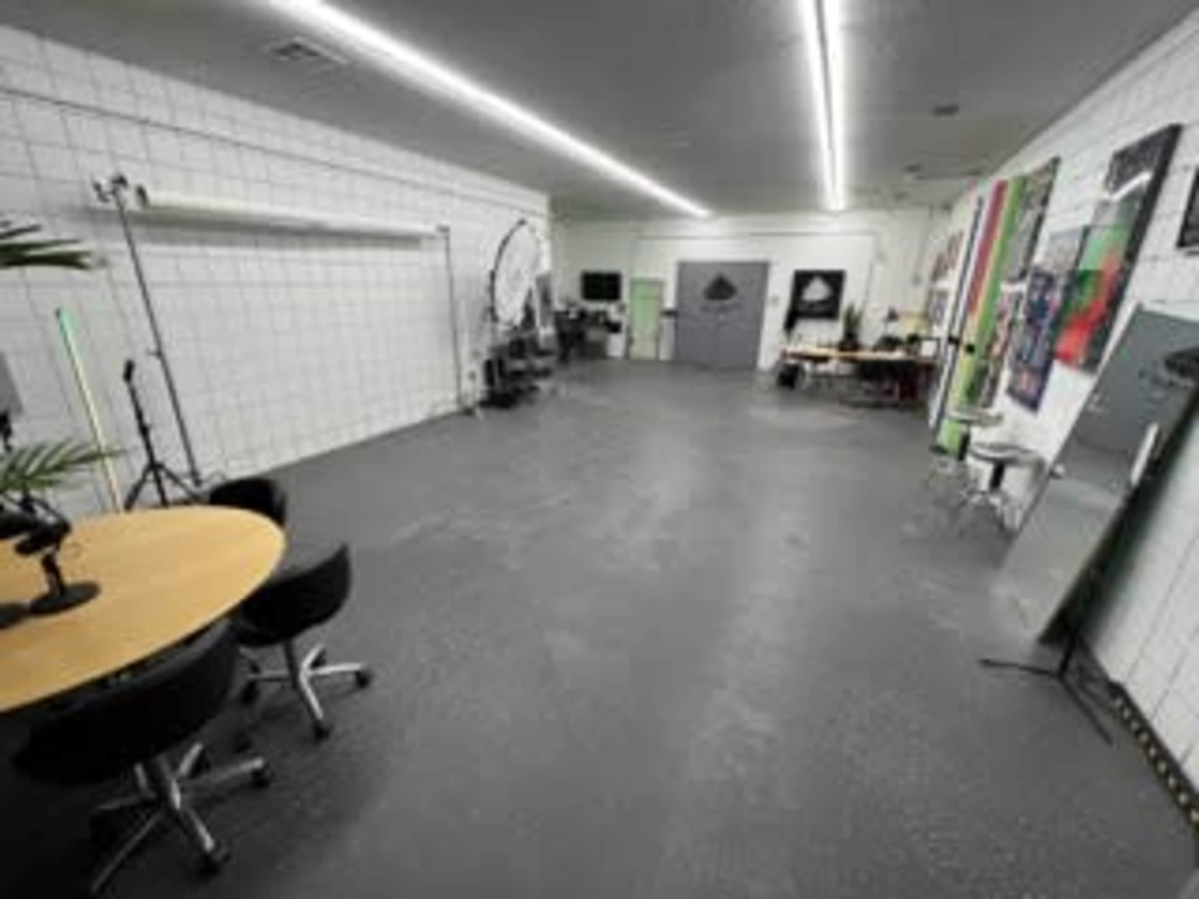A spacious studio with a gray floor, white tiled walls, a round table with black chairs, and various equipment and artwork along the walls.