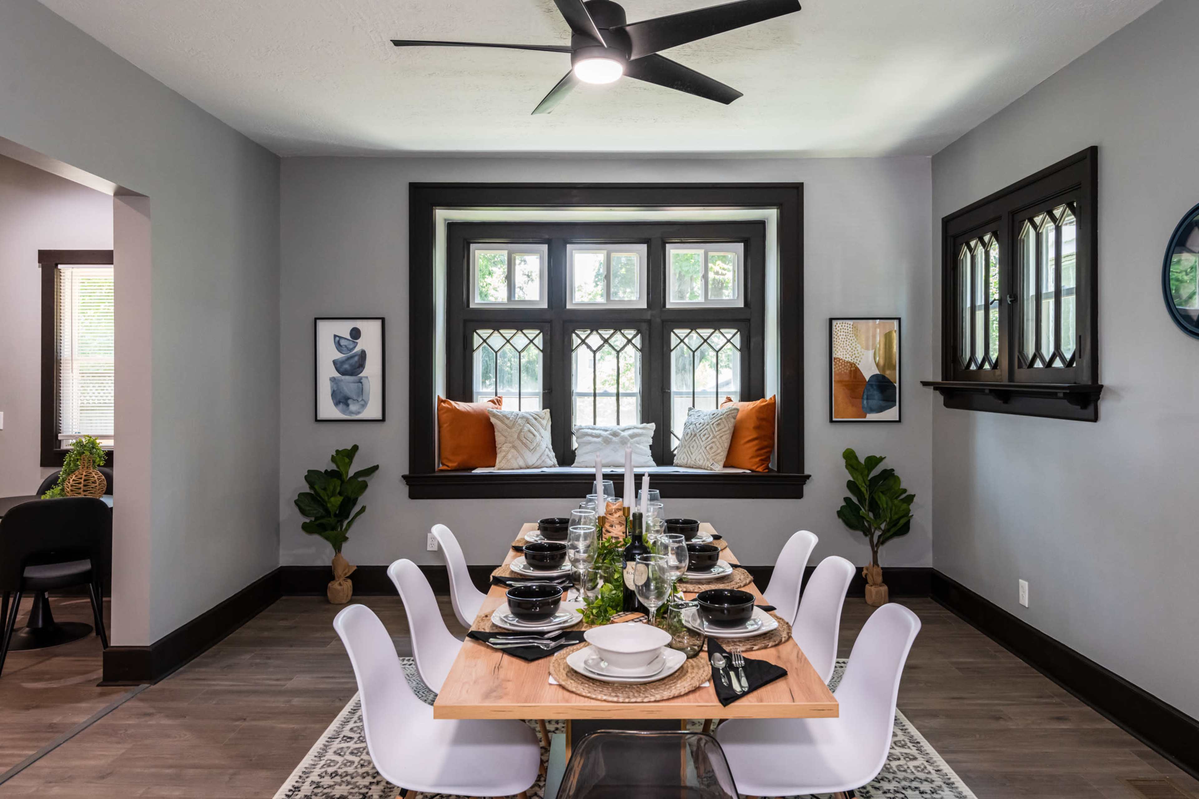 A dining area features a table set for eight, with a large window showcasing decorative cushions, framed artwork, and potted plants.