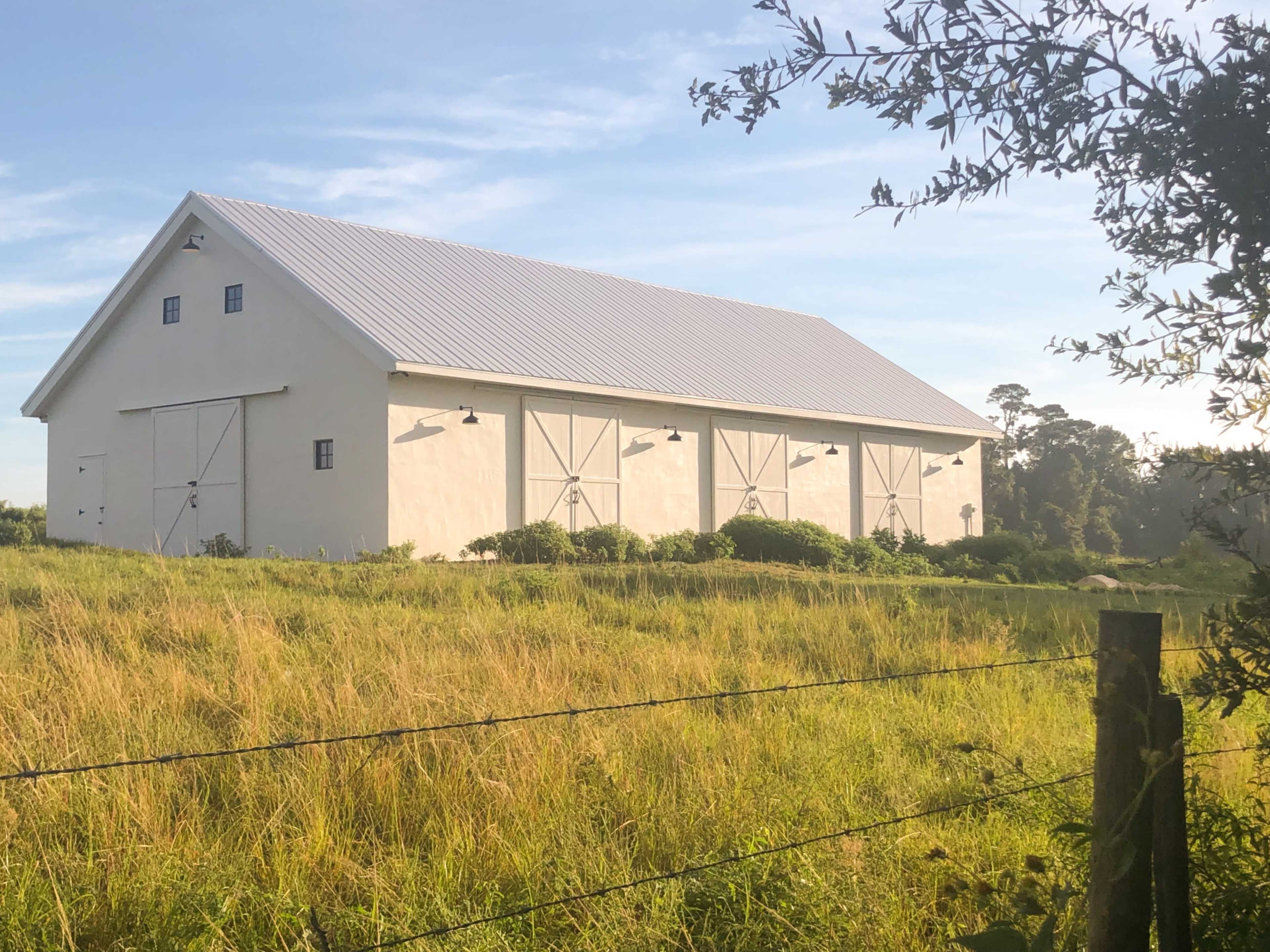 A large white barn with a metal roof sits in a grassy field, surrounded by trees.