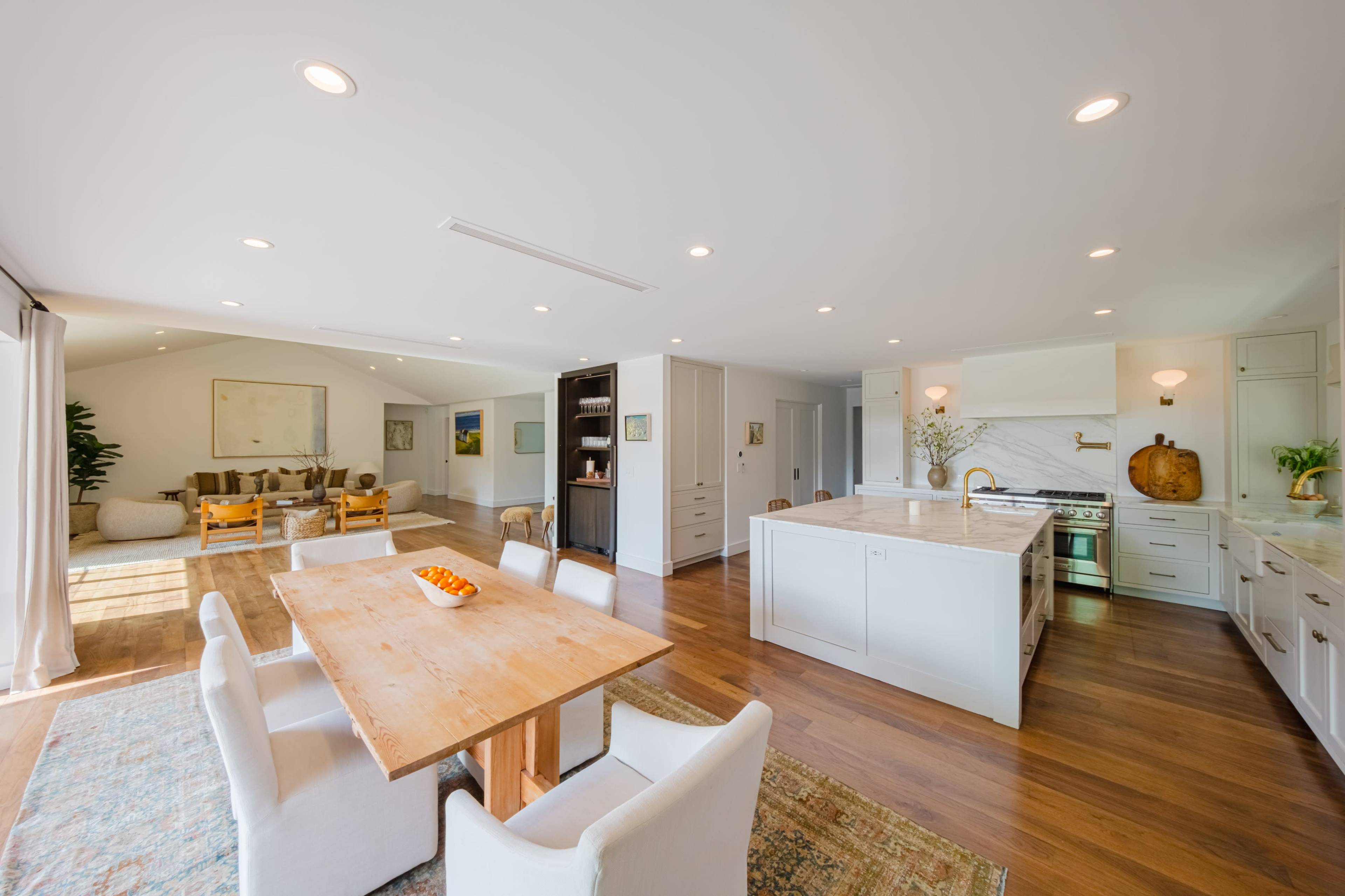 A modern open-concept living space features a dining area with a wooden table, surrounded by white chairs, adjacent to a kitchen with sleek cabinetry and appliances.
