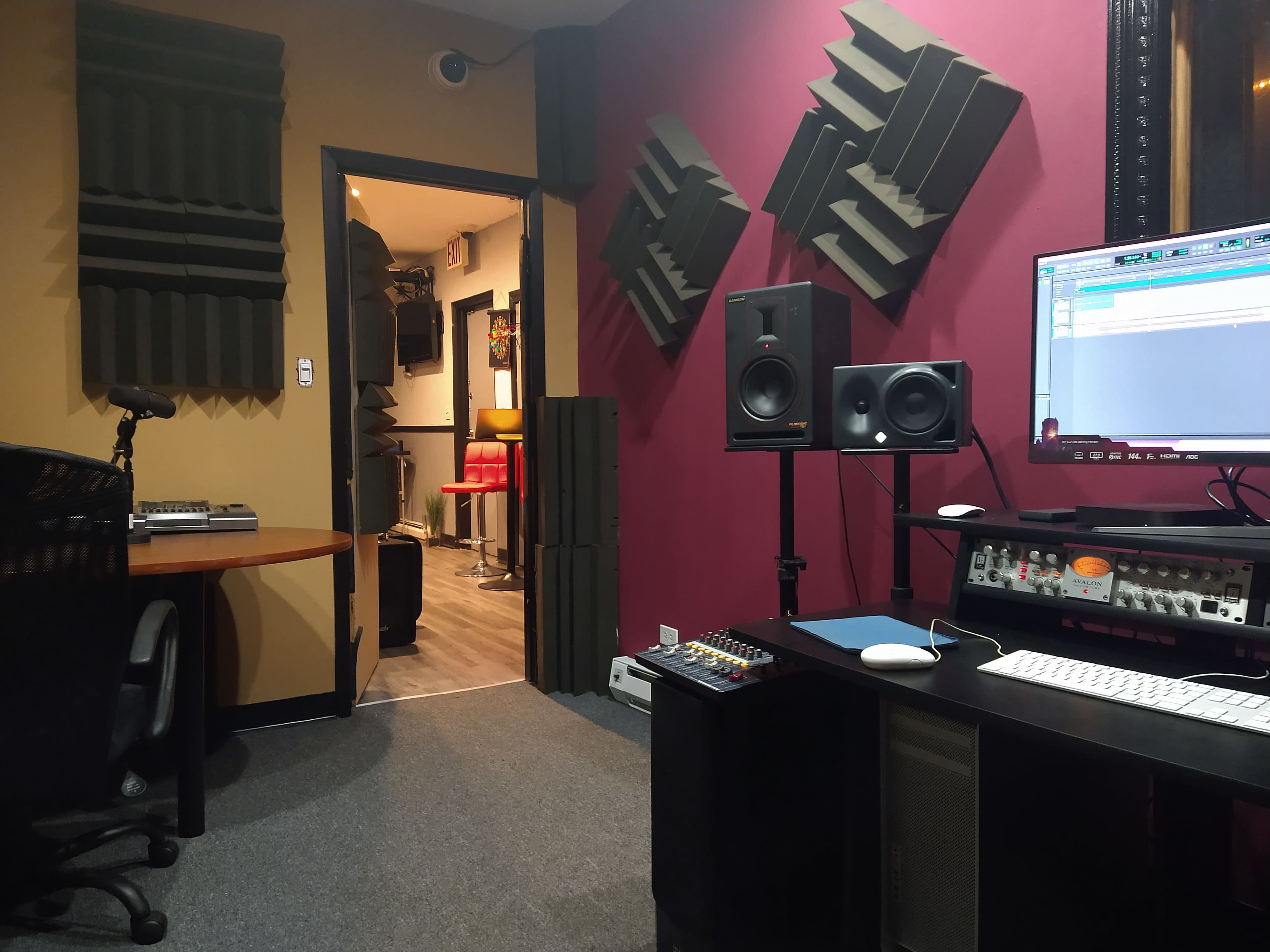 A recording studio featuring soundproofing panels on the walls, a computer workstation with speakers, and an open doorway leading to another room.