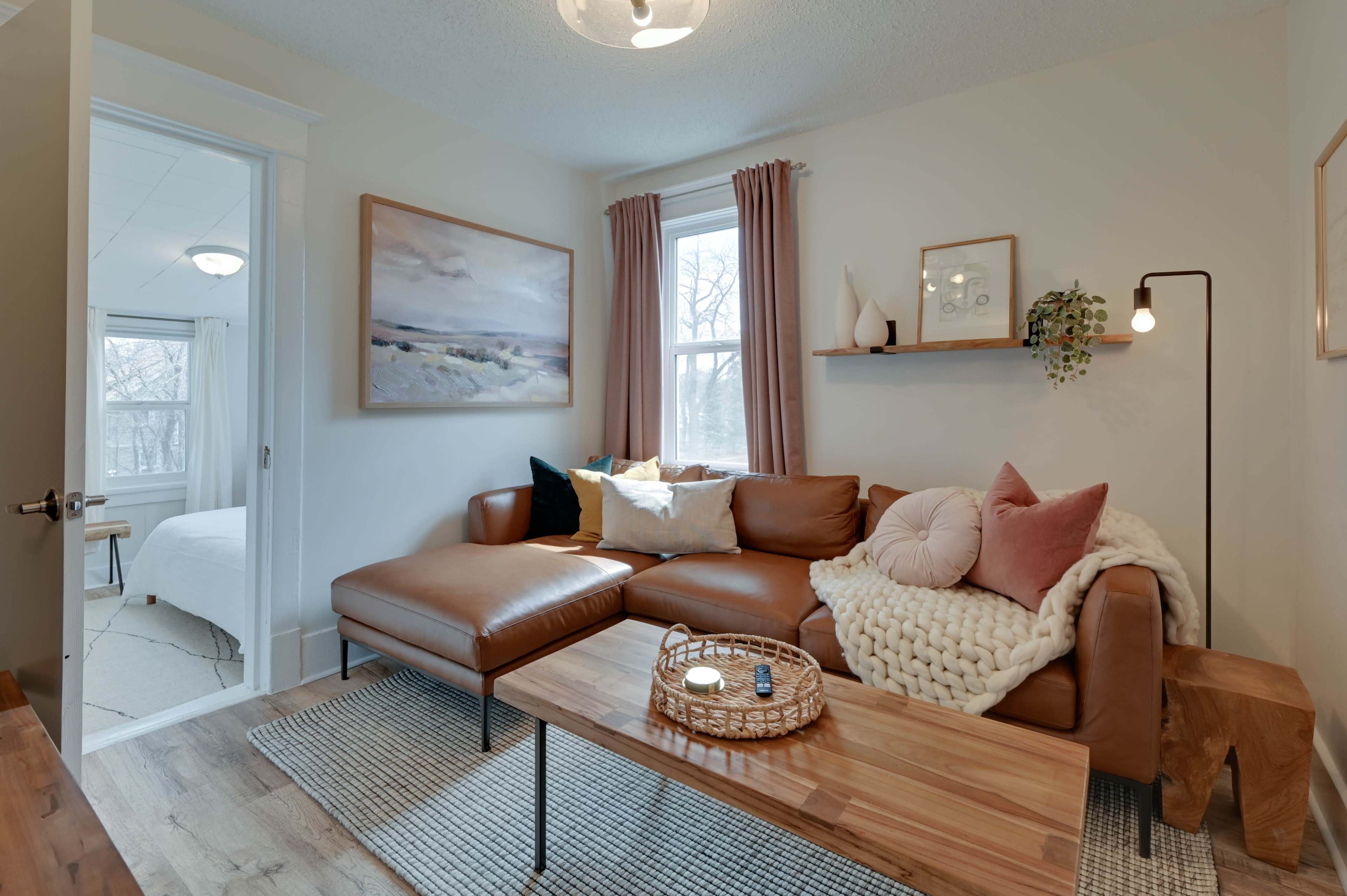 A cozy living room features a brown leather sectional sofa, a wooden coffee table, and a decorative wall shelf, with natural light streaming through a window.