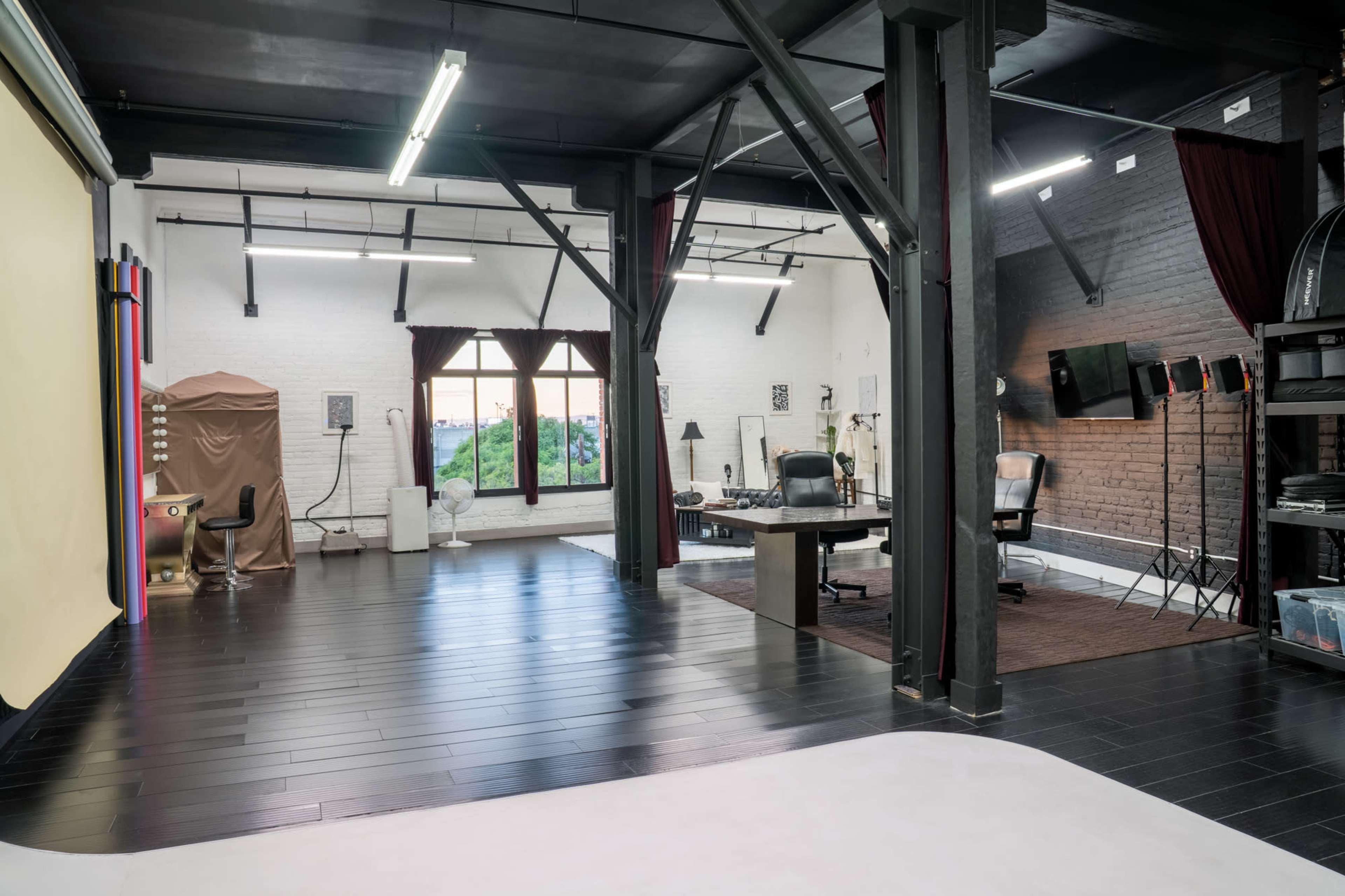 The image shows a spacious studio with wooden beams, large windows, and a variety of furniture including a desk and chairs, along with a backdrop setup in one corner.