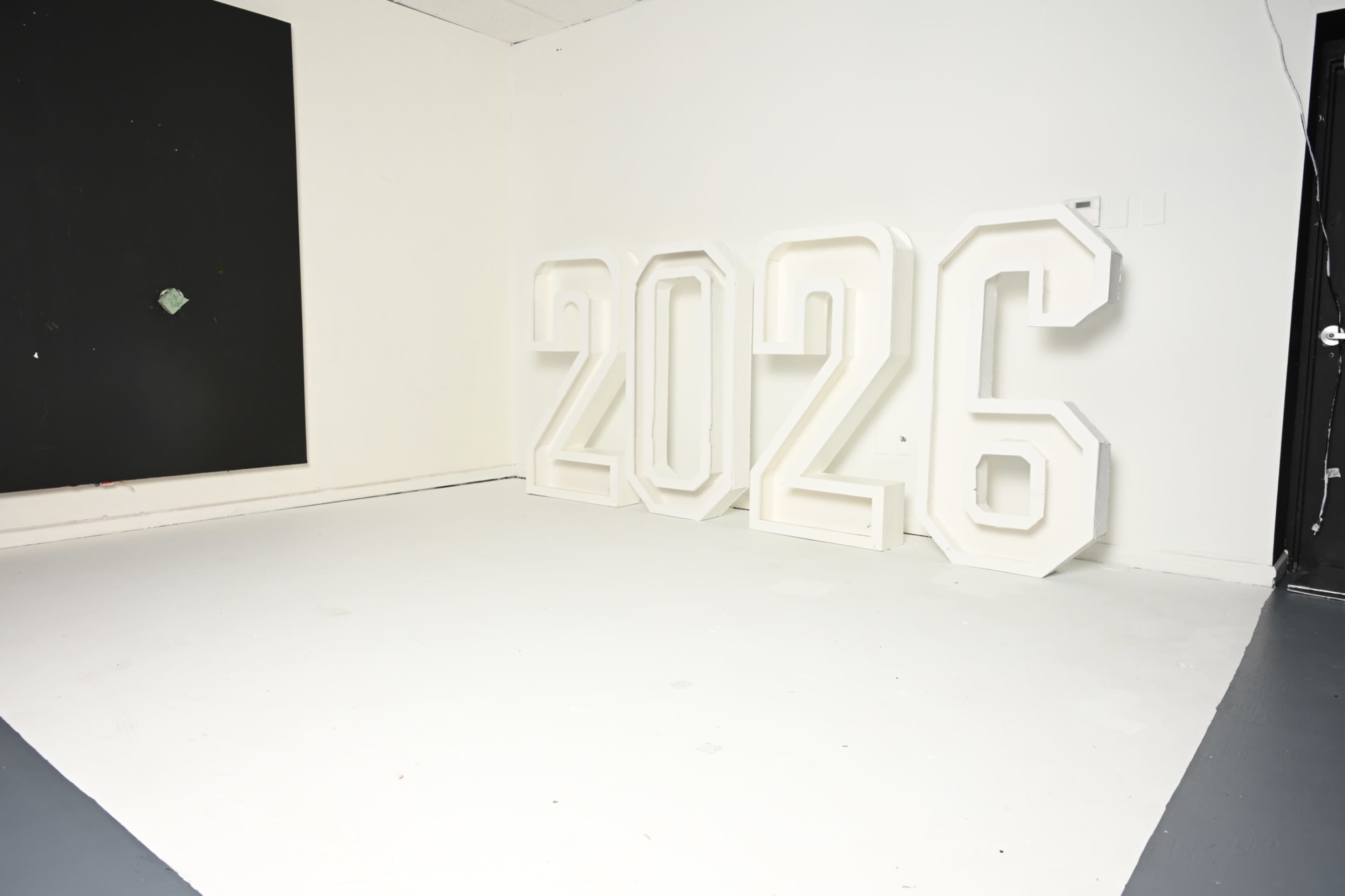 The image shows large, white numeral sculptures spelling out "2026" against a blank wall in a minimalistic room.