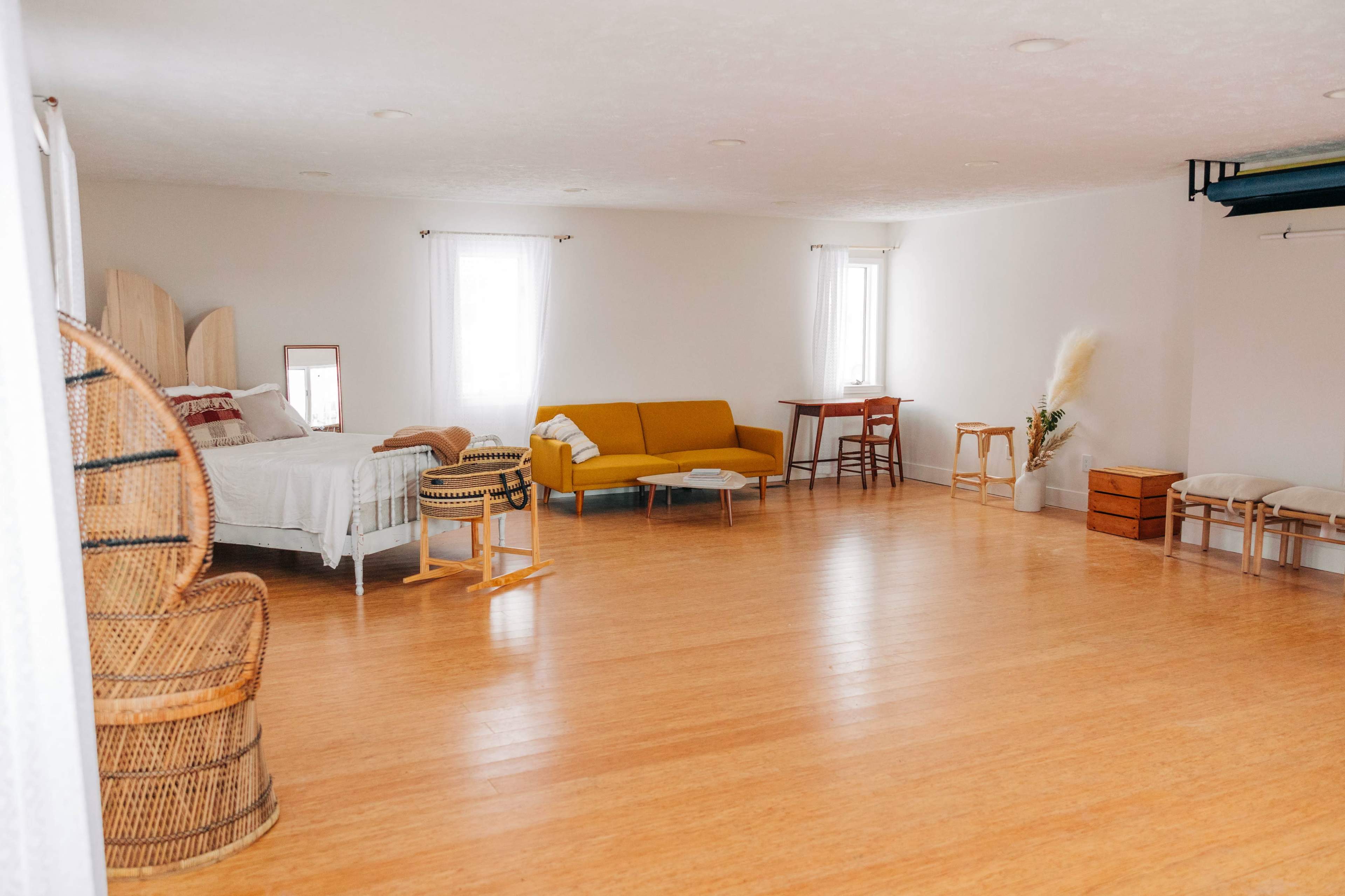 A spacious, minimalist room with a bed, a yellow sofa, a wooden table, and various pieces of furniture arranged on a light wooden floor.