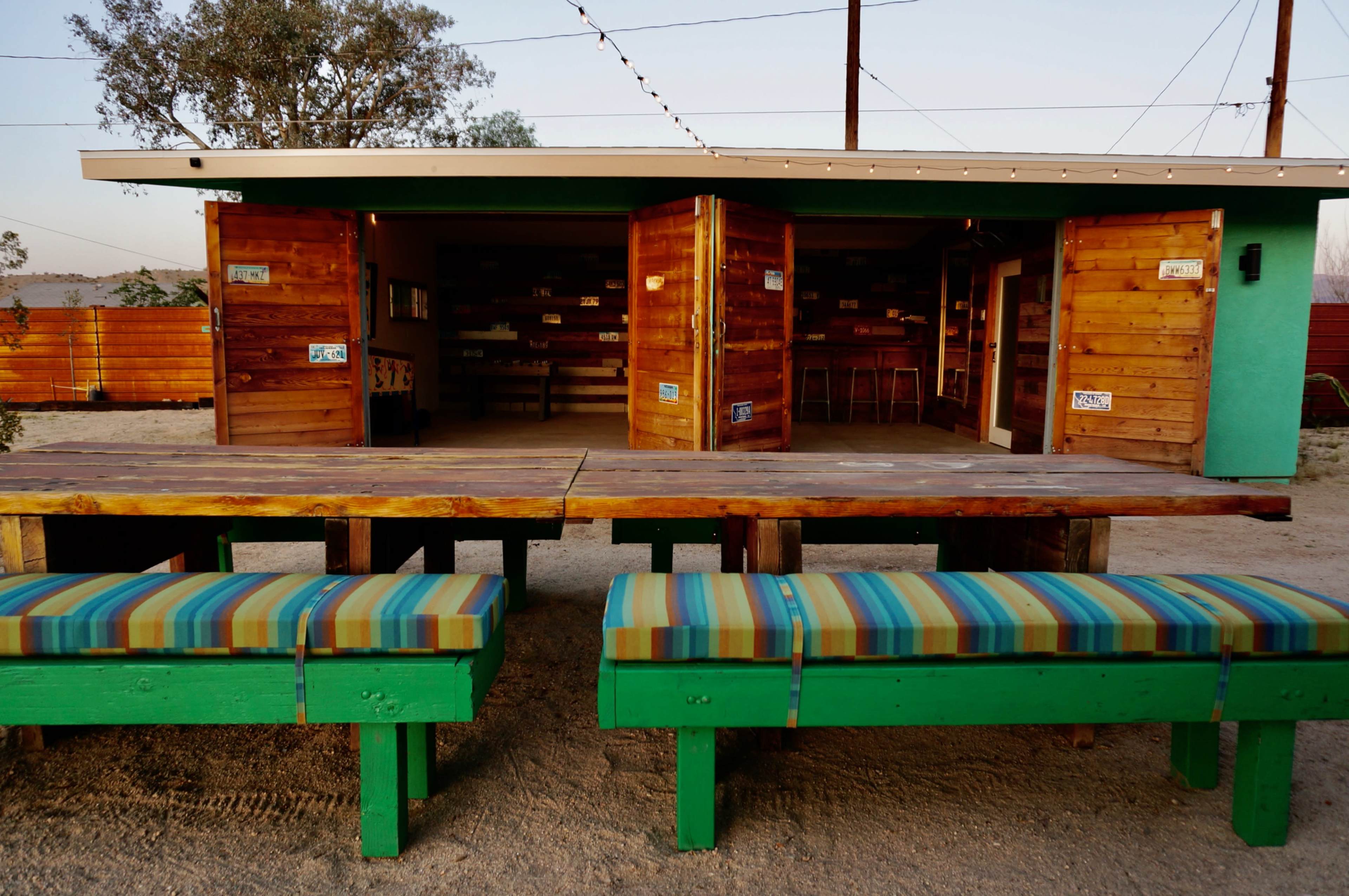 The image shows a wooden building with open doors and a long bench with colorful cushions in front, situated in a desert area.