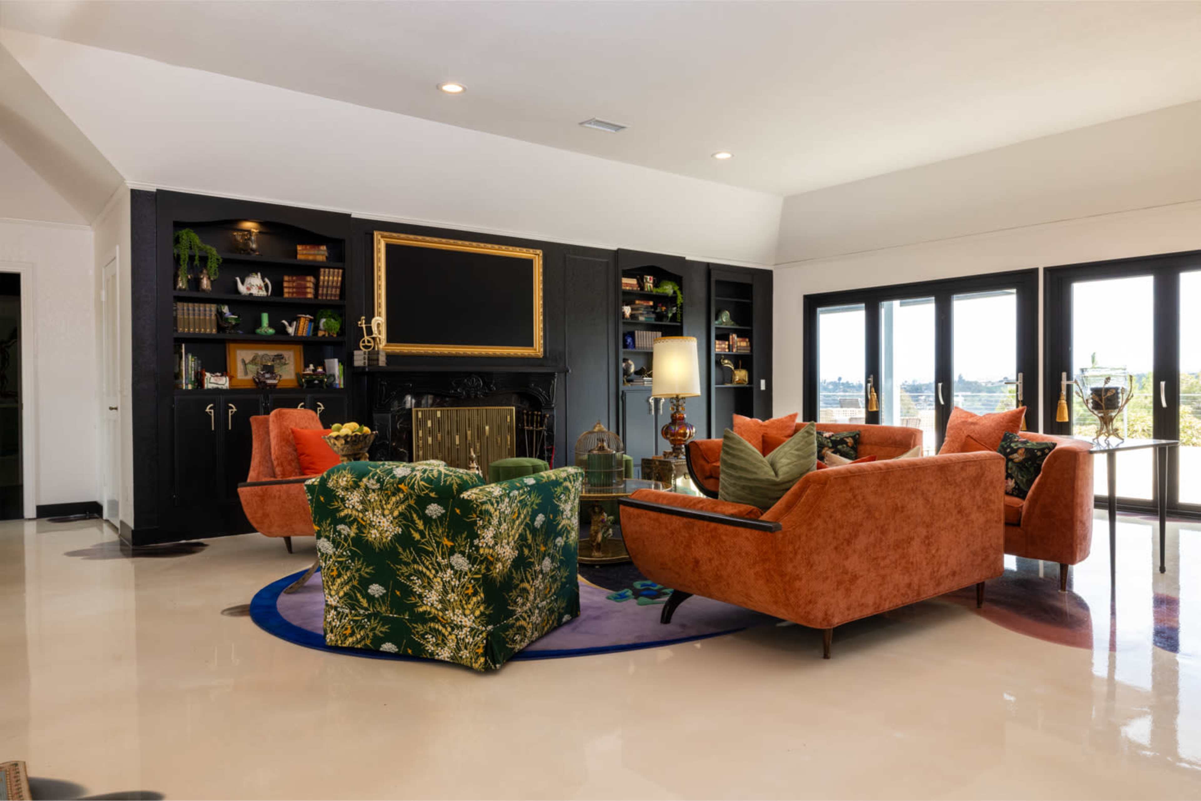 The scene features a modern living room with two orange armchairs and a green patterned chair arranged around a circular rug, facing a black entertainment unit with a large screen.
