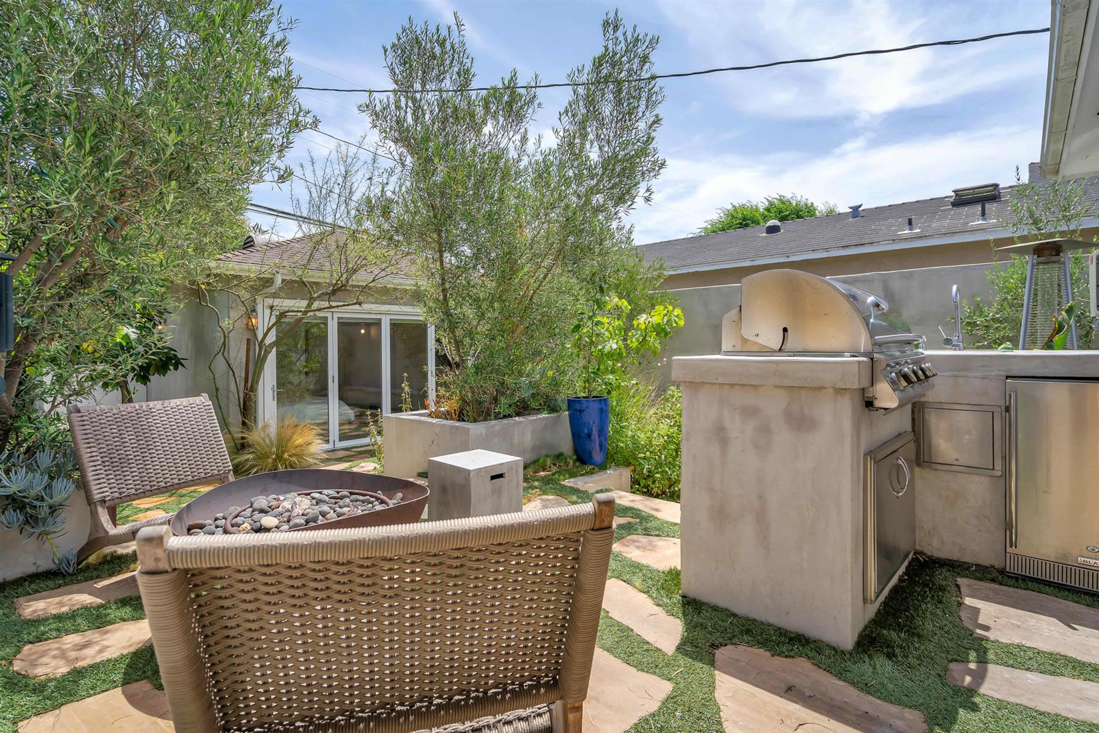 The image shows a patio area with two woven chairs, a fire pit with stones, a barbecue grill, and various plants around a stone pathway.