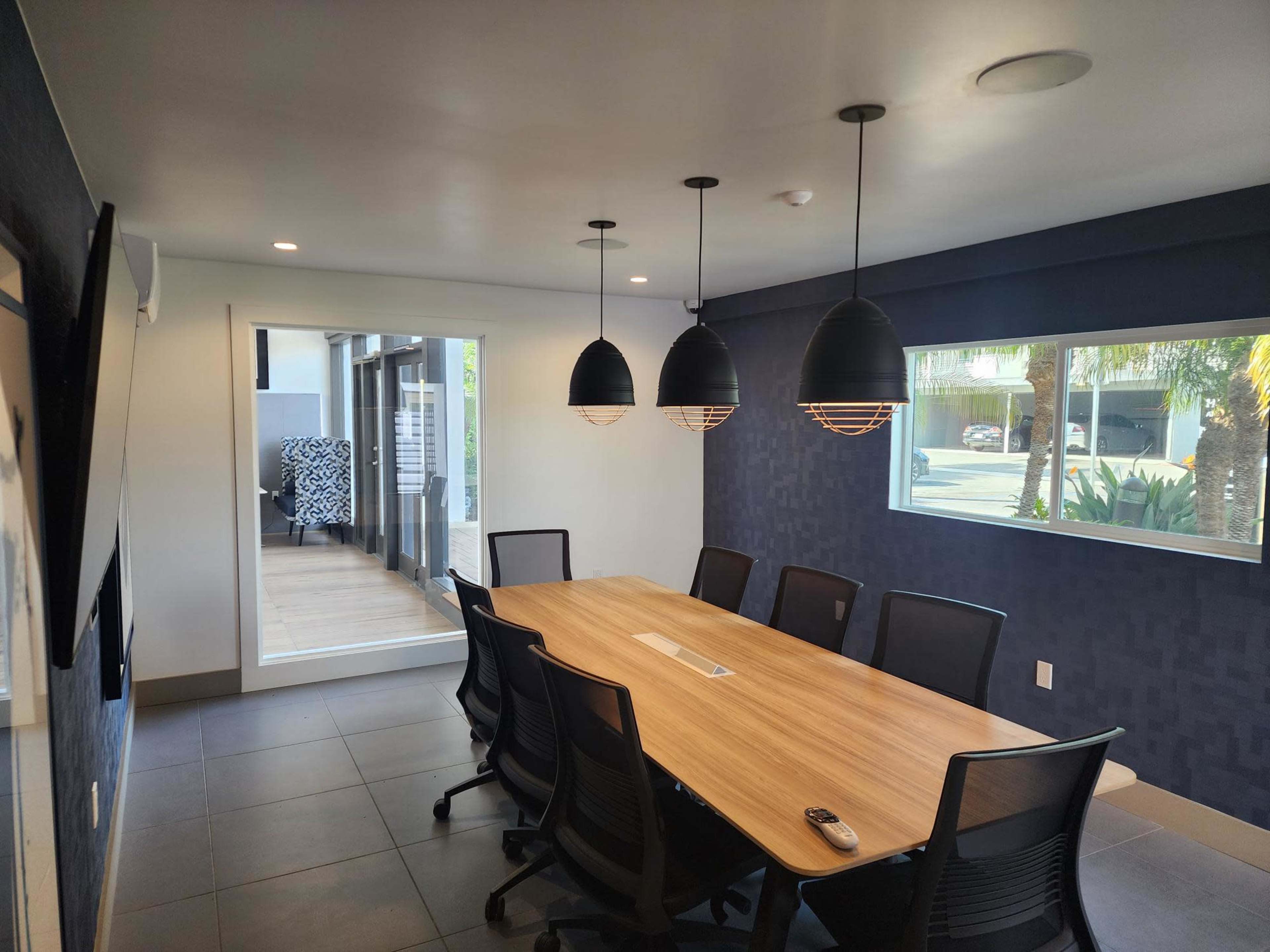 A modern conference room with a large wooden table surrounded by black chairs, illuminated by three pendant lights.