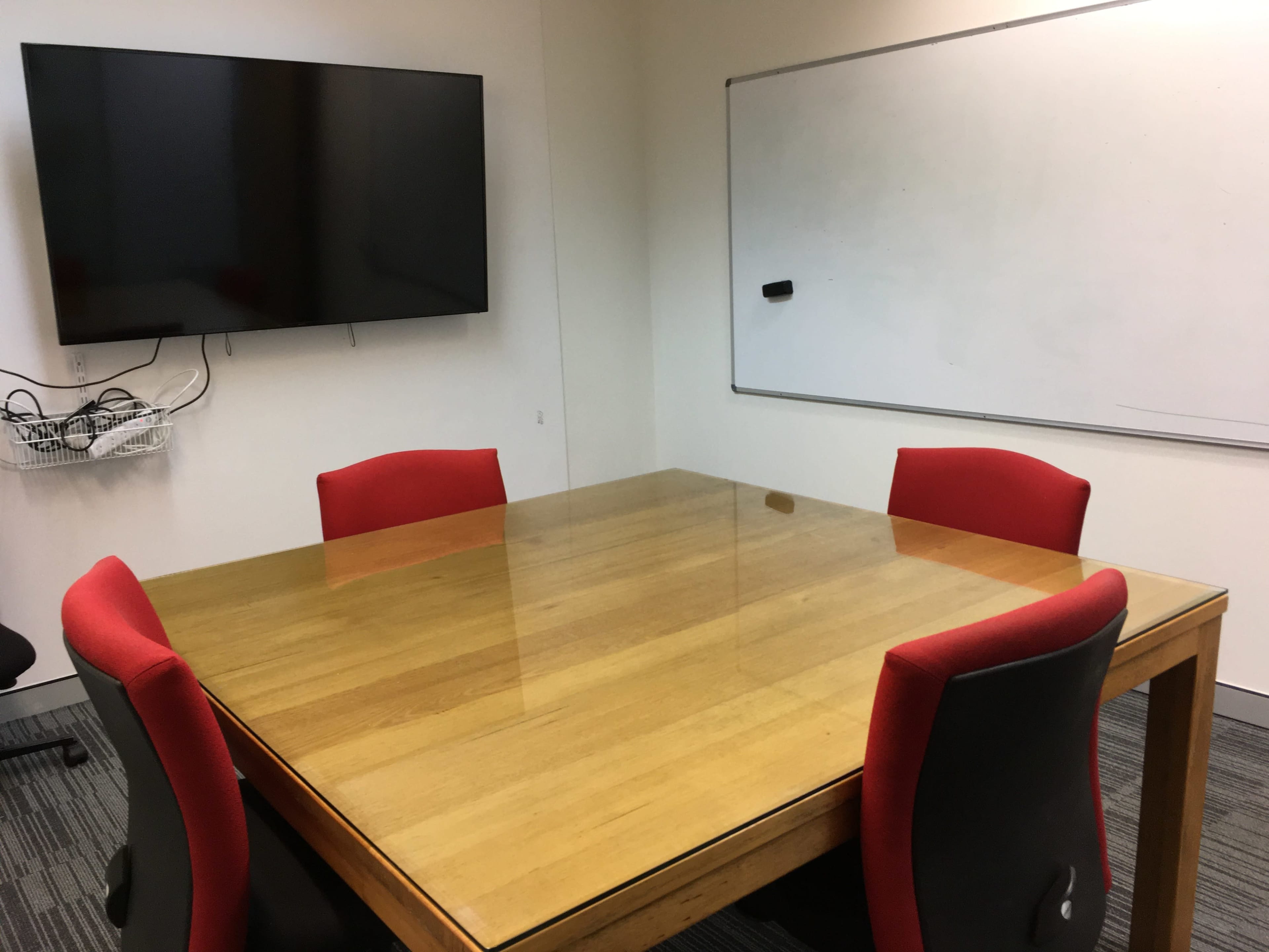 A conference room features a large wooden table surrounded by four red chairs, with a wall-mounted television and a whiteboard nearby.