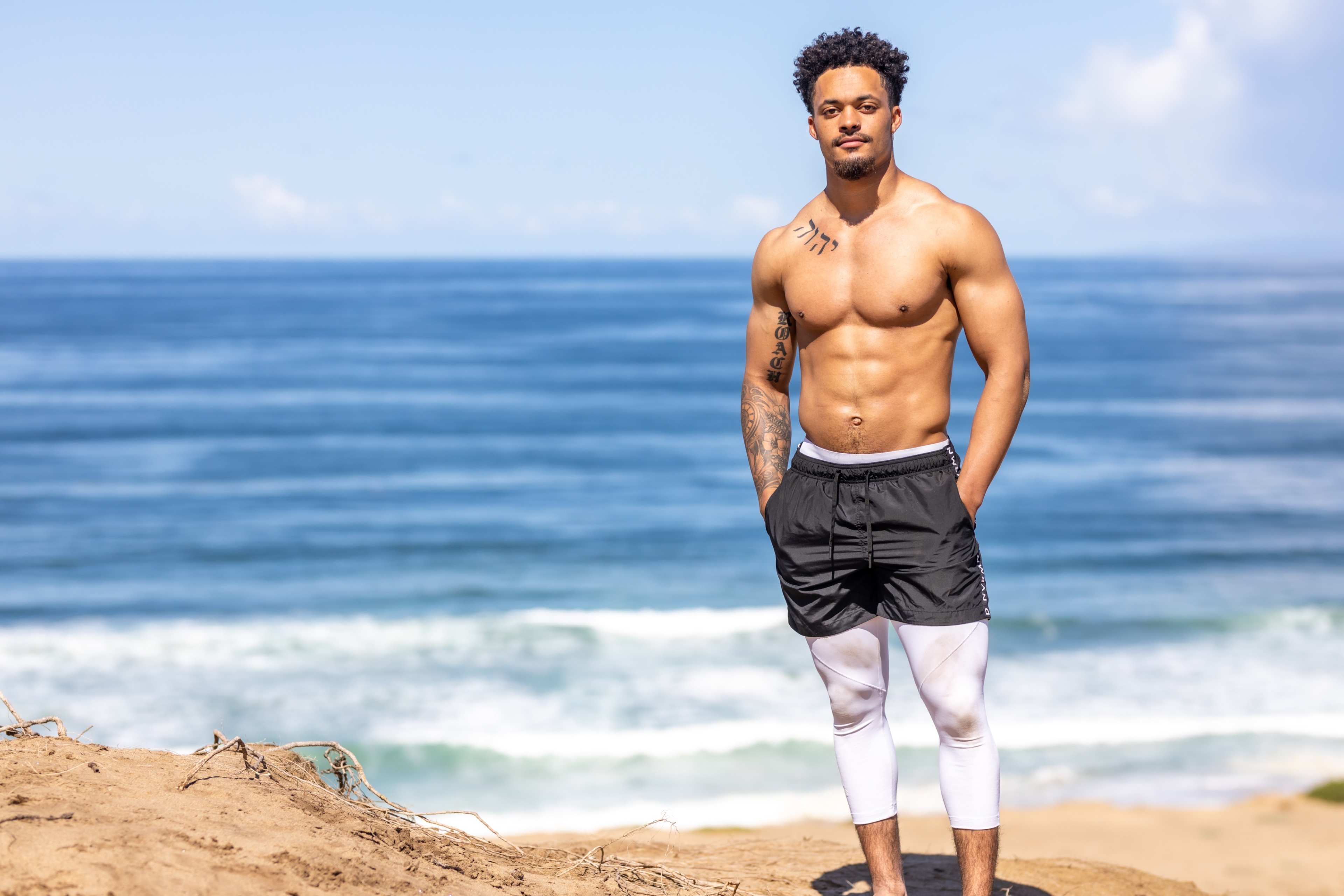 A shirtless man with tattoos stands on a sandy shoreline overlooking the ocean.