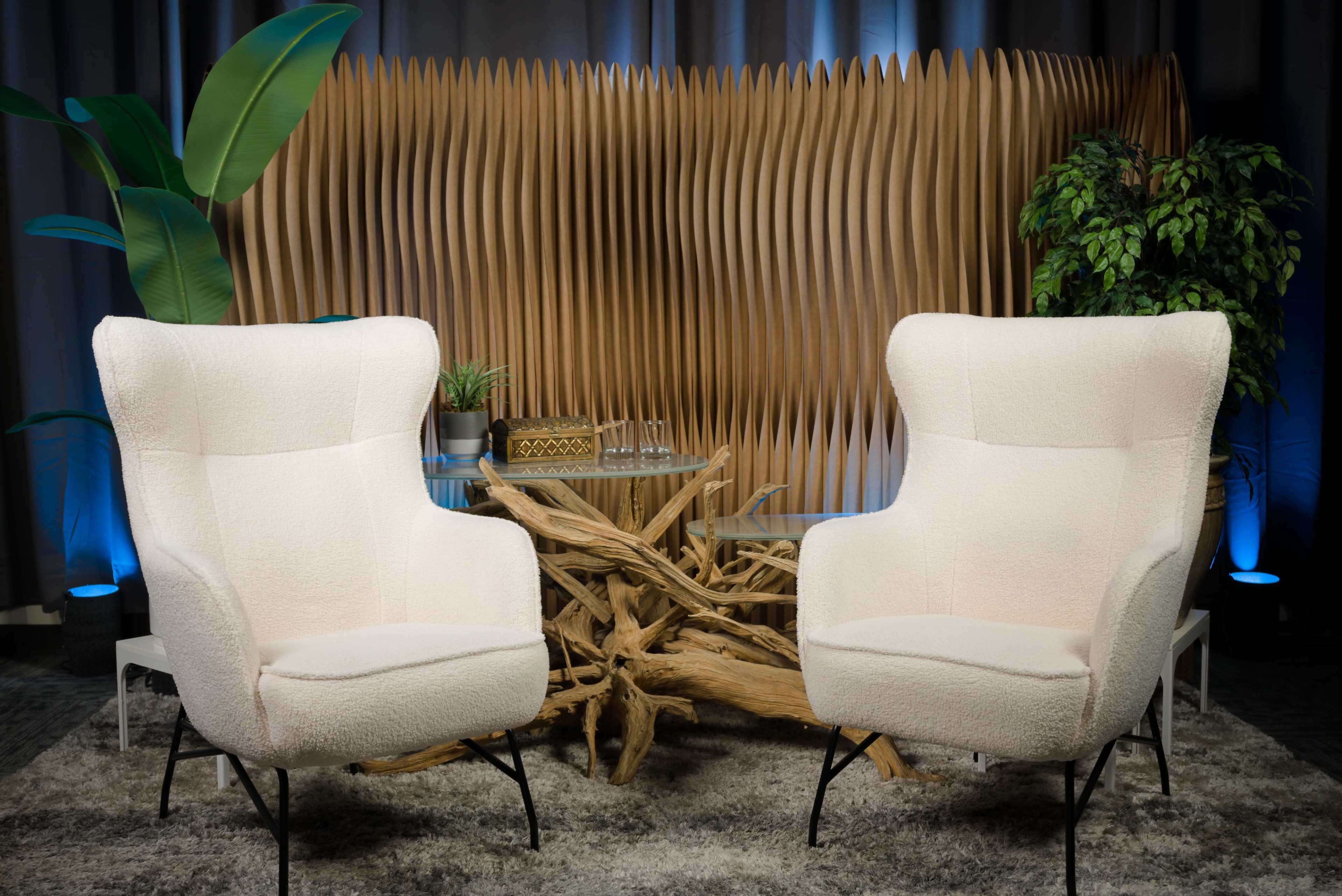 The image features two white armchairs facing each other in a modern setting, with a wooden artistic background and decorative plants.