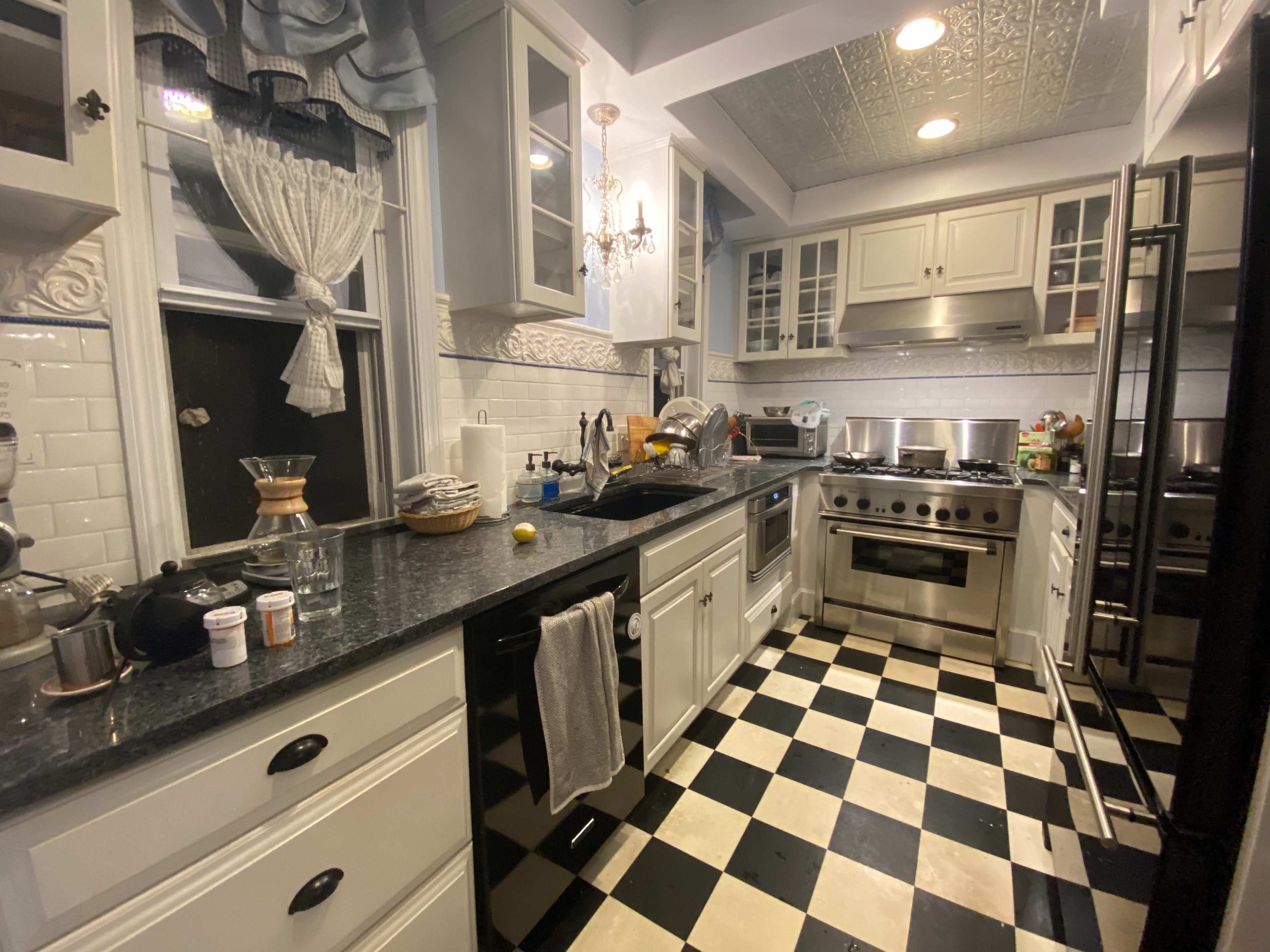 The image shows a kitchen with black and white checkered flooring, stainless steel appliances, and white cabinetry, featuring a mixture of kitchen utensils and clutter on the countertop.