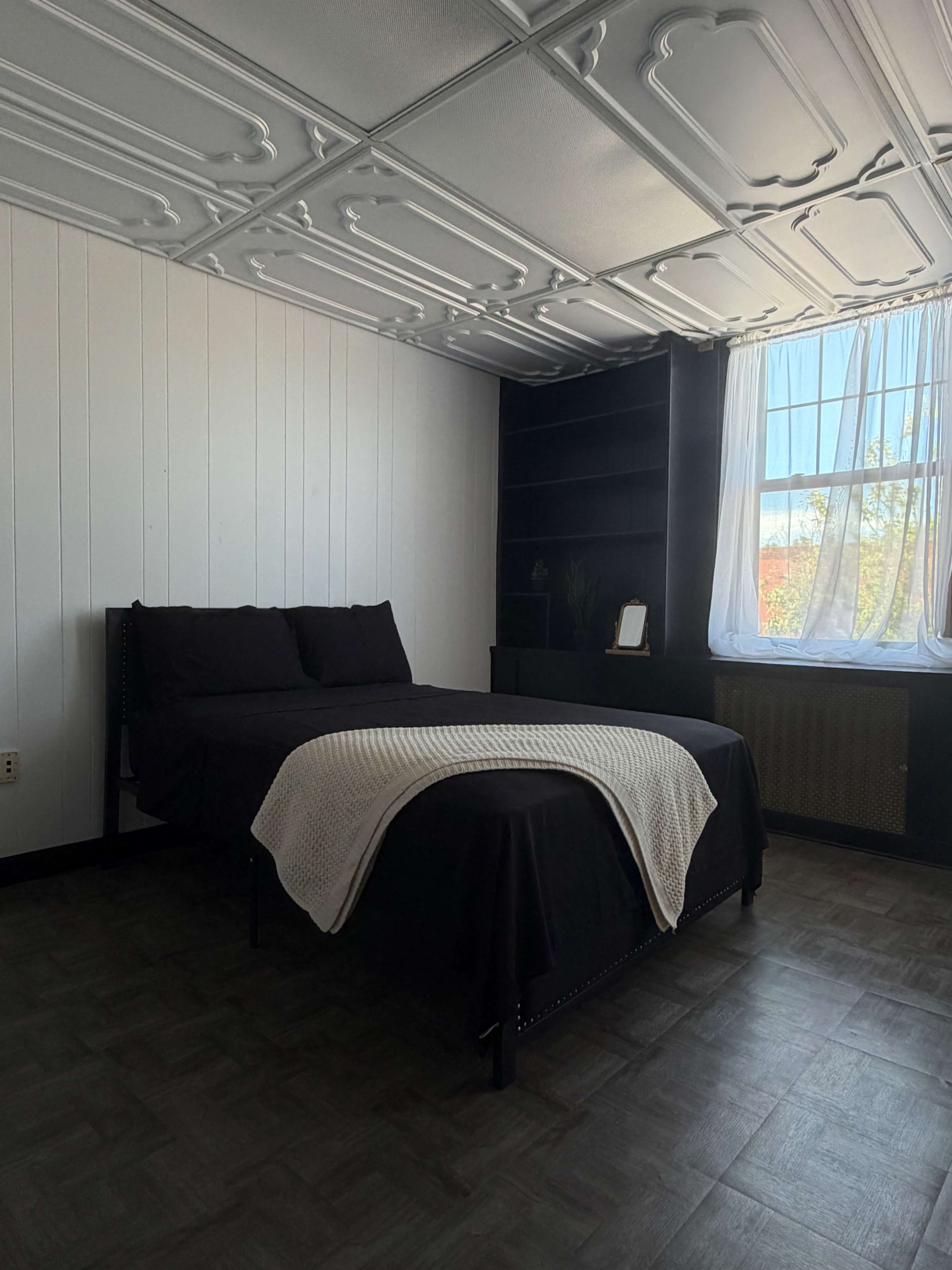 The image shows a minimalist bedroom with a black bed, a white throw blanket, and a window letting in natural light.