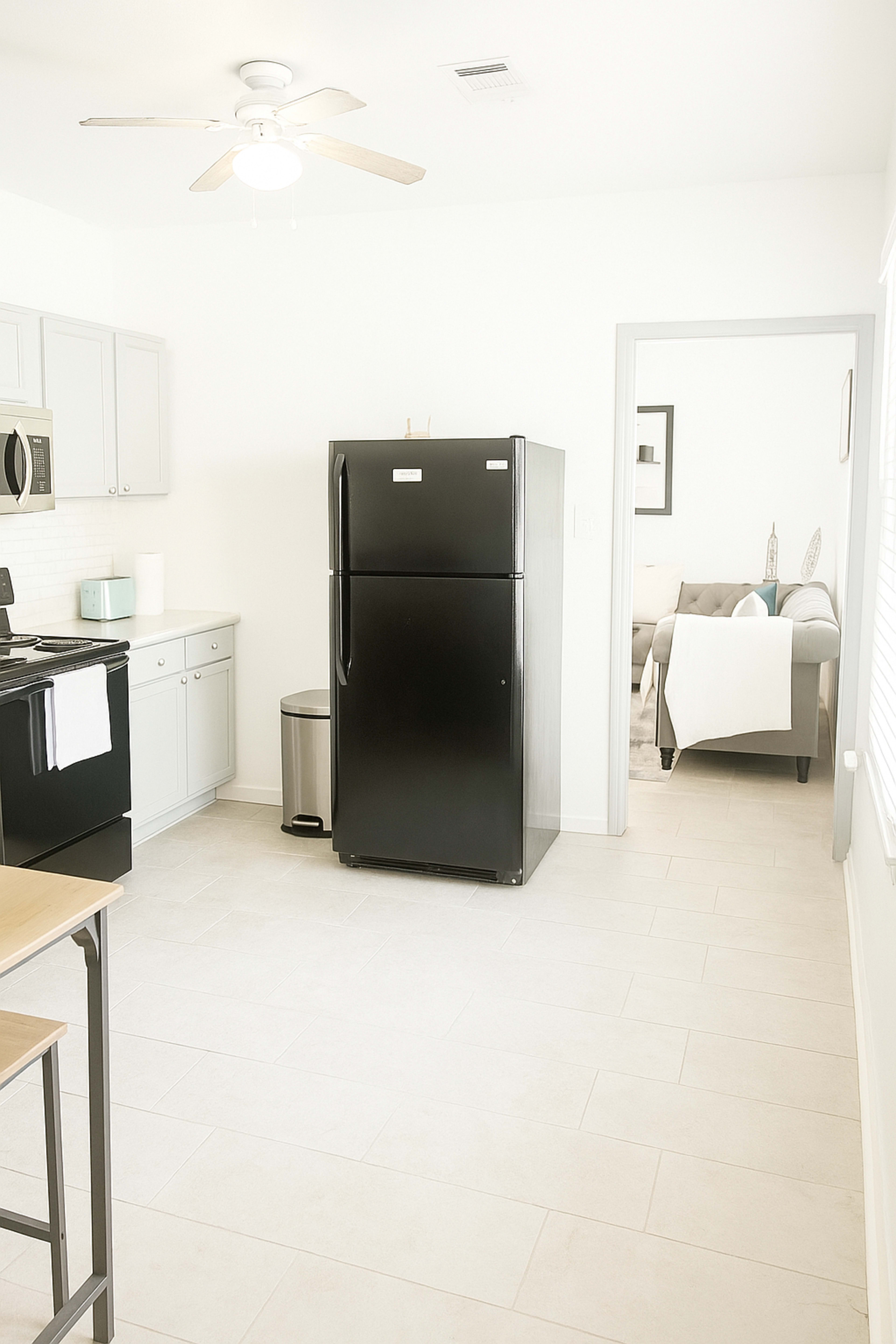 The image shows a modern kitchen featuring a black refrigerator, light gray cabinets, a stove, and a dining area with a visible living room in the background.
