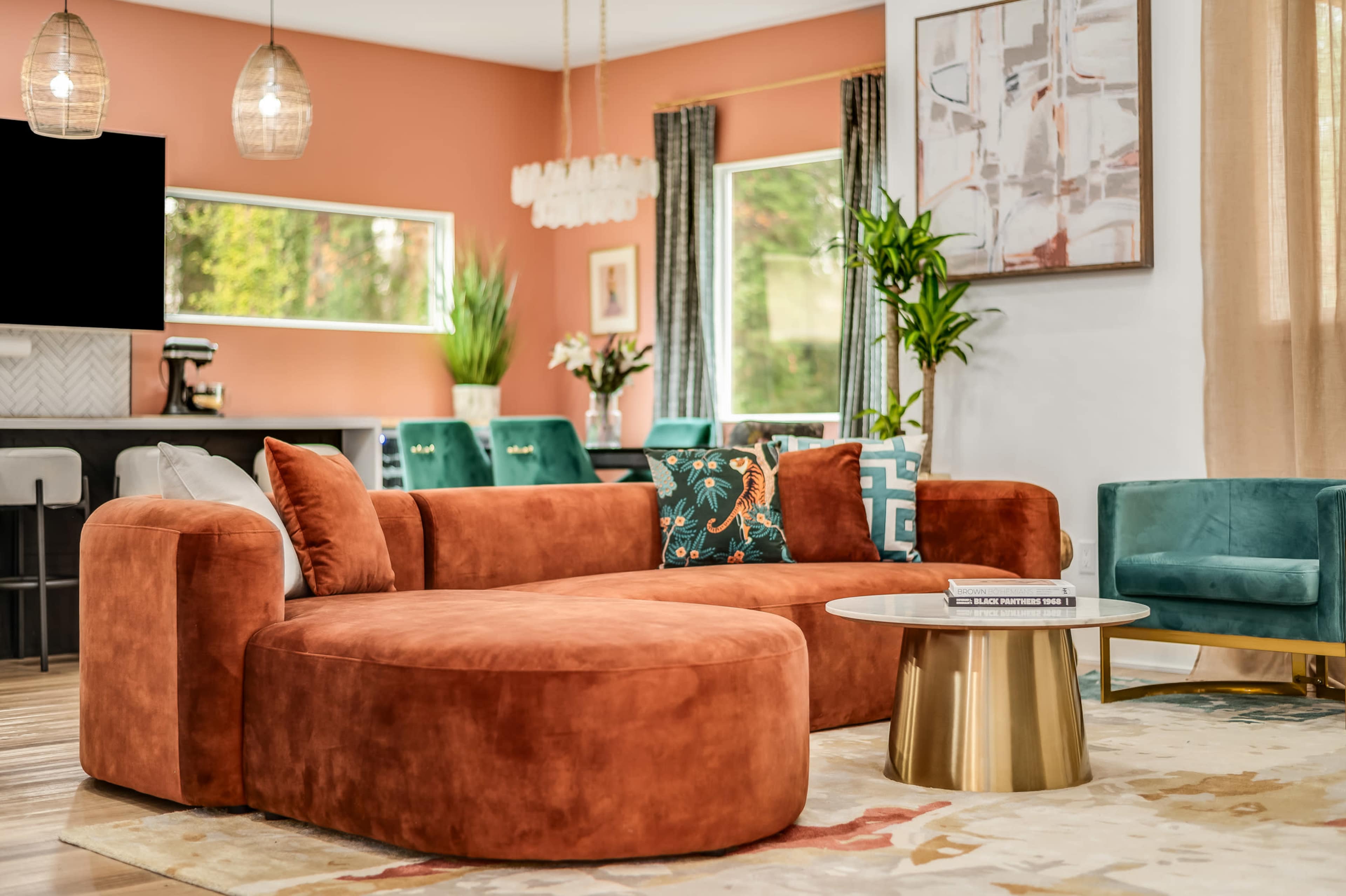 The image shows a stylish living room featuring an orange sectional sofa, a circular coffee table, and decorative plants, with a modern dining area in the background.